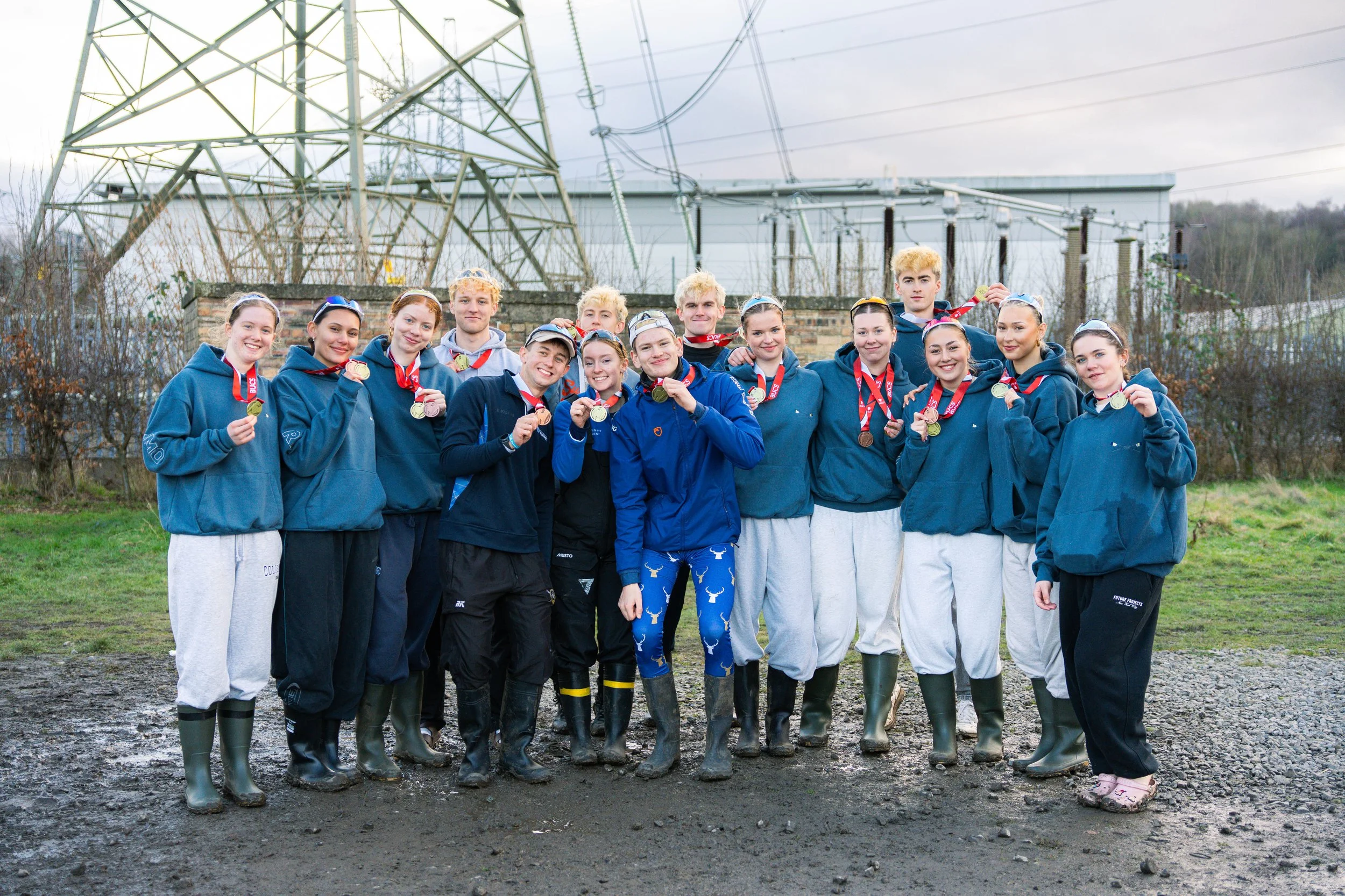 A group of young people wearing blue hoodies and rain boots, standing outdoors on a muddy ground, holding medals and smiling. In the background are electrical transmission towers and overcast sky.