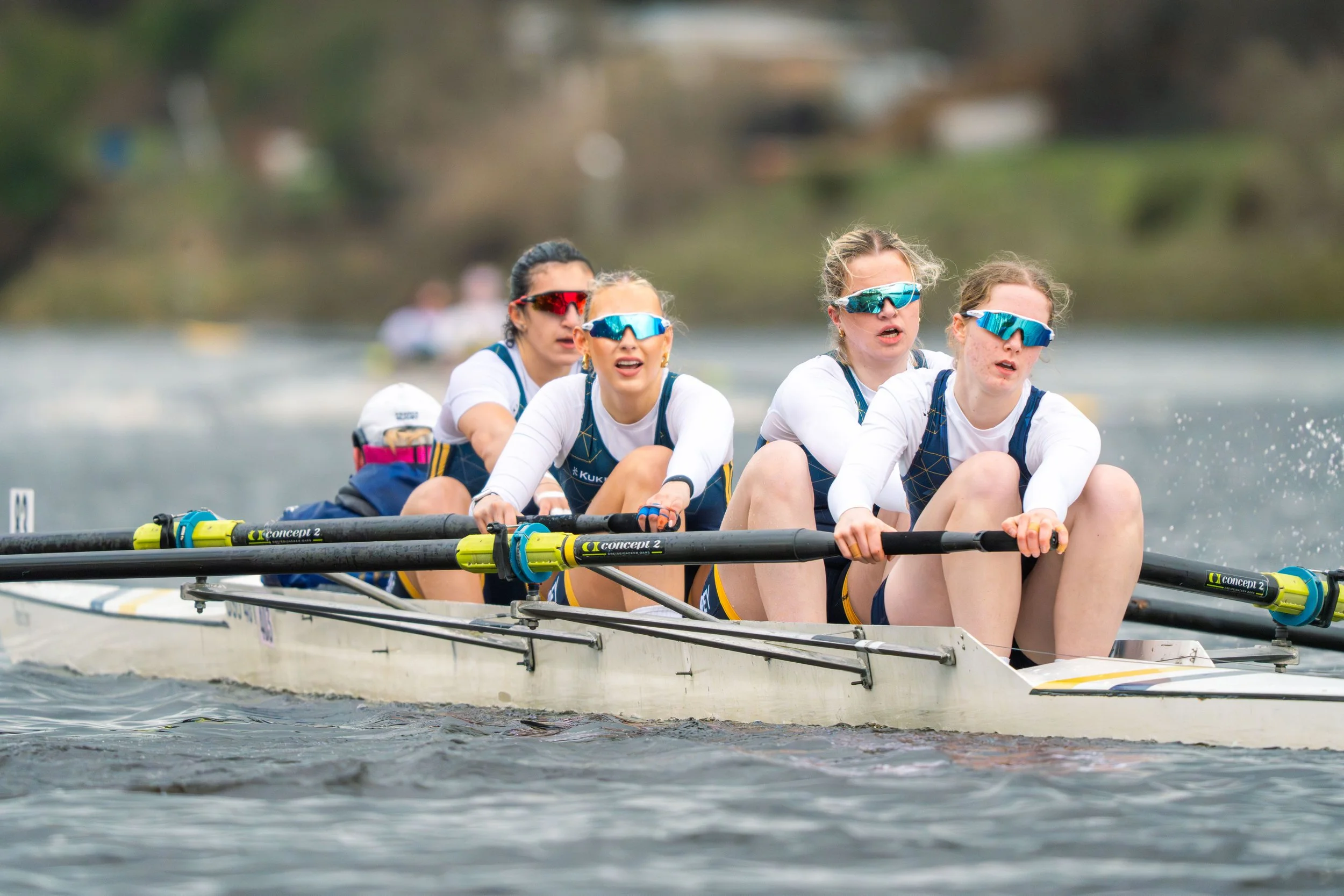 Four female rowers wearing sunglasses and team uniforms rowing a boat on the water.