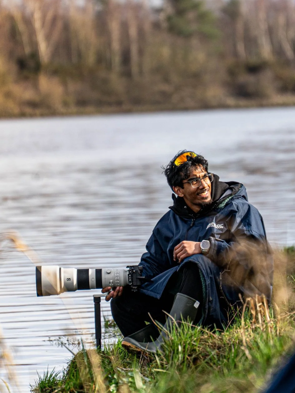 A man sitting on the grass near a lake with a camera, smiling and looking to his right. He is wearing glasses, a black jacket, and yellow sunglasses on his head.