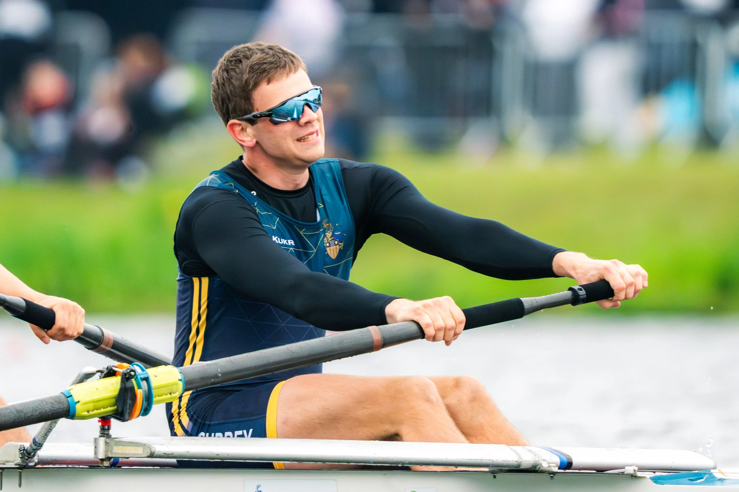 A male athlete in a blue and black rowing uniform wearing sunglasses, participating in a rowing sport on a body of water, with other rowers in the background.