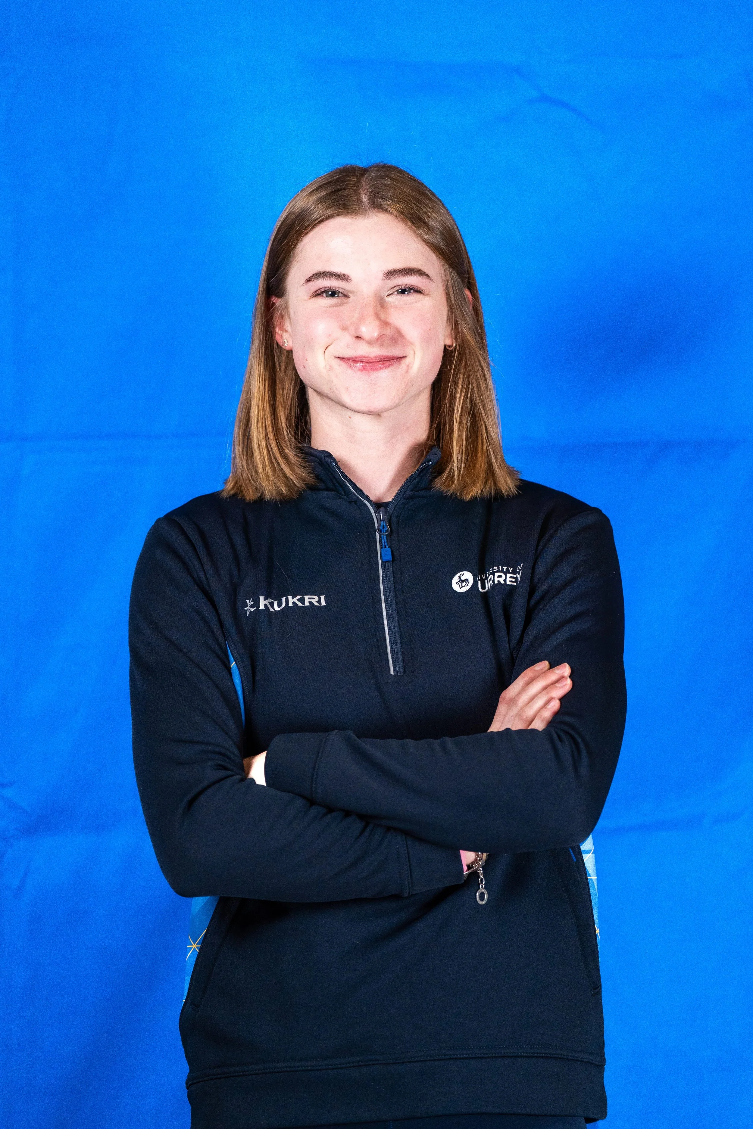 A young woman with brown hair smiling and crossing her arms, wearing a black jacket with logos, standing against a blue background.