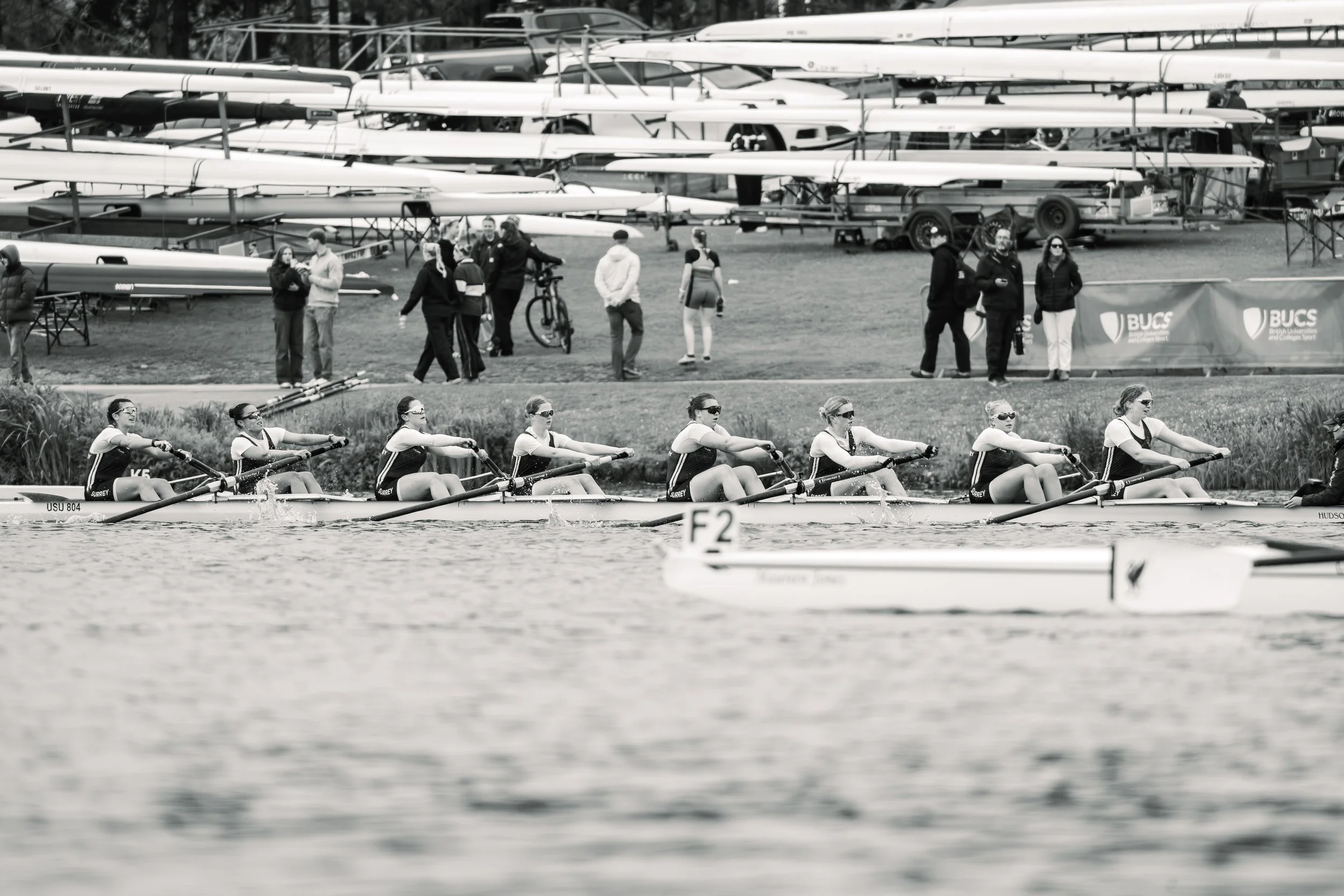 A group of female rowers in a boat on a river, with people and boats on racks in the background, indicating a rowing event or practice session.