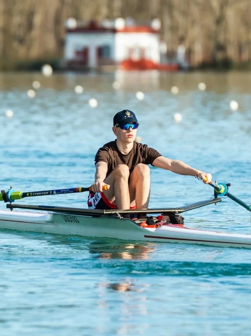 A person in a kayak on the water, wearing sunglasses and a baseball cap, with a house in the background.