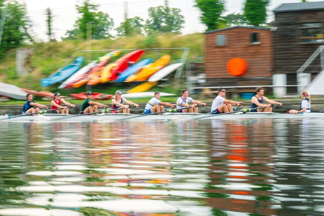 A team of eight people rowing a boat on a lake with colorful kayaks stored on a dock in the background