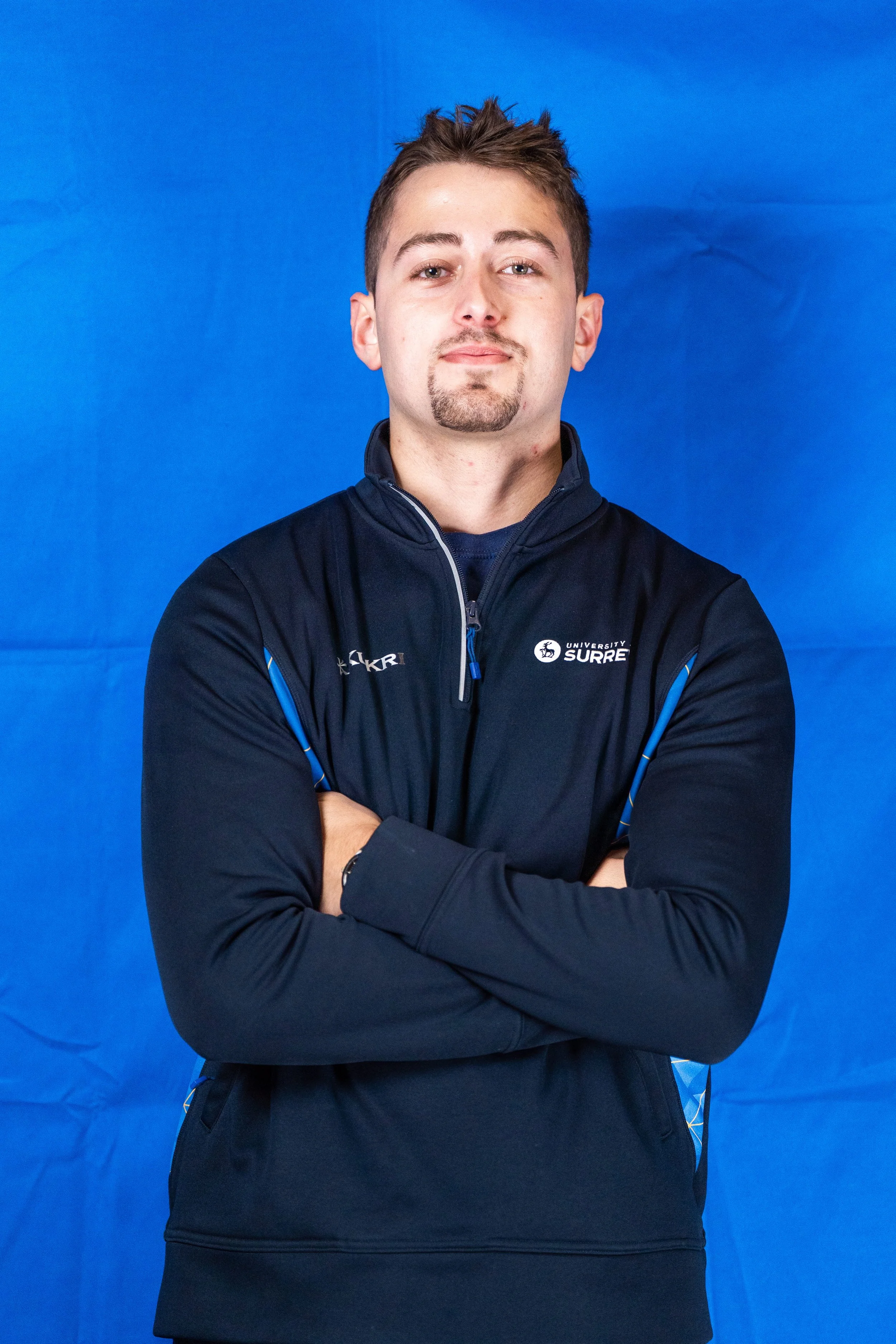 Portrait of a young man with short brown hair, a goatee, wearing a navy blue athletic jacket with logos, standing with arms crossed against a solid blue background.