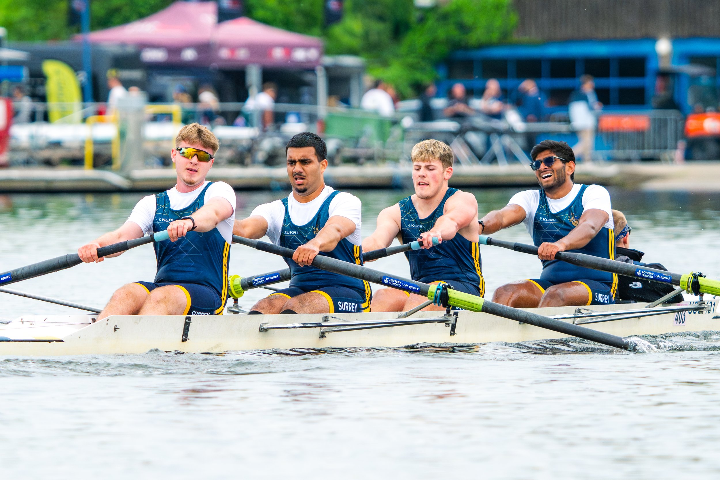 Four male rowers on a racing boat competing in a rowing competition on a river, with spectators and tents in the background.