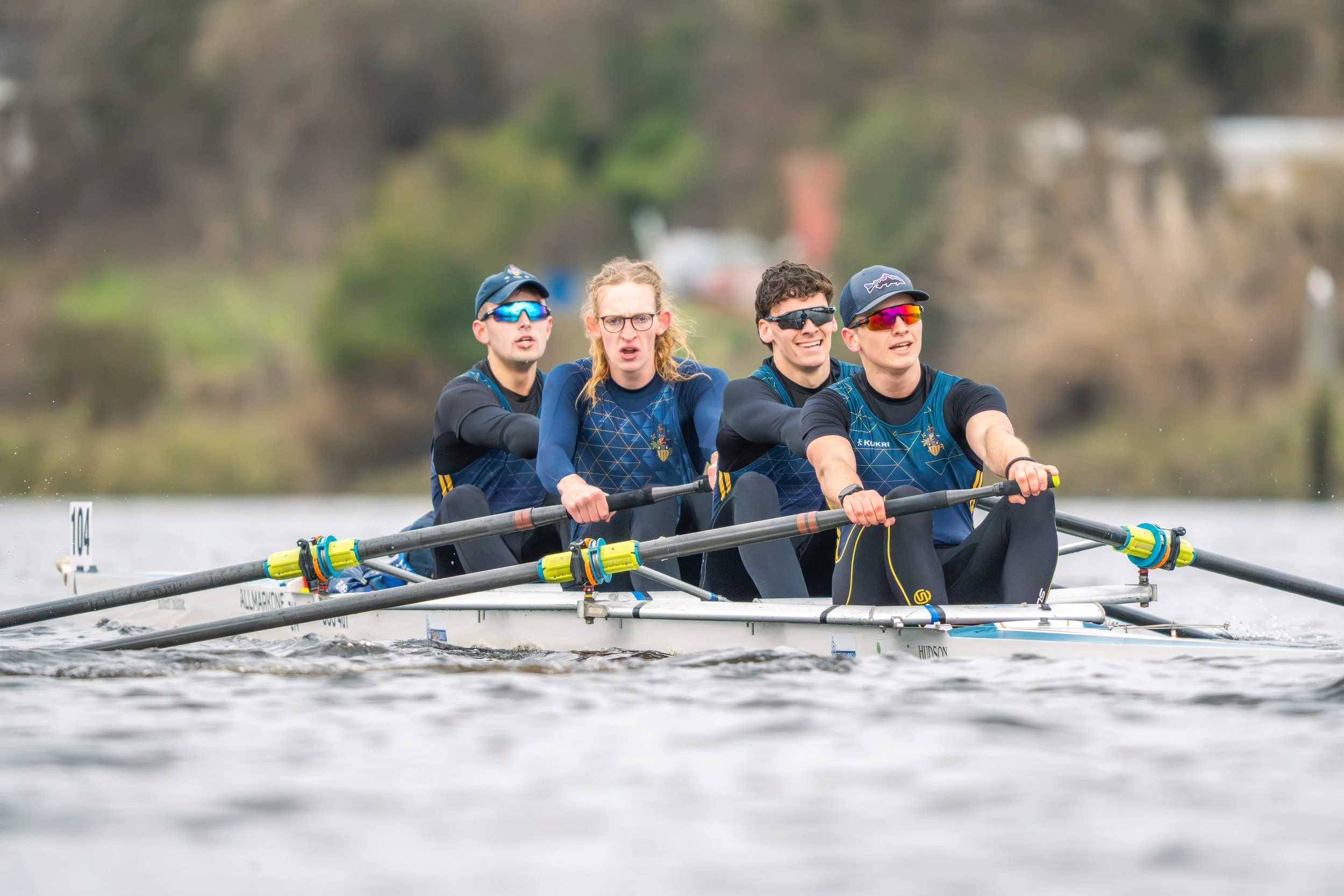 A group of four rowers in a boat on the water, wearing athletic gear and sunglasses, with a blurred natural background.