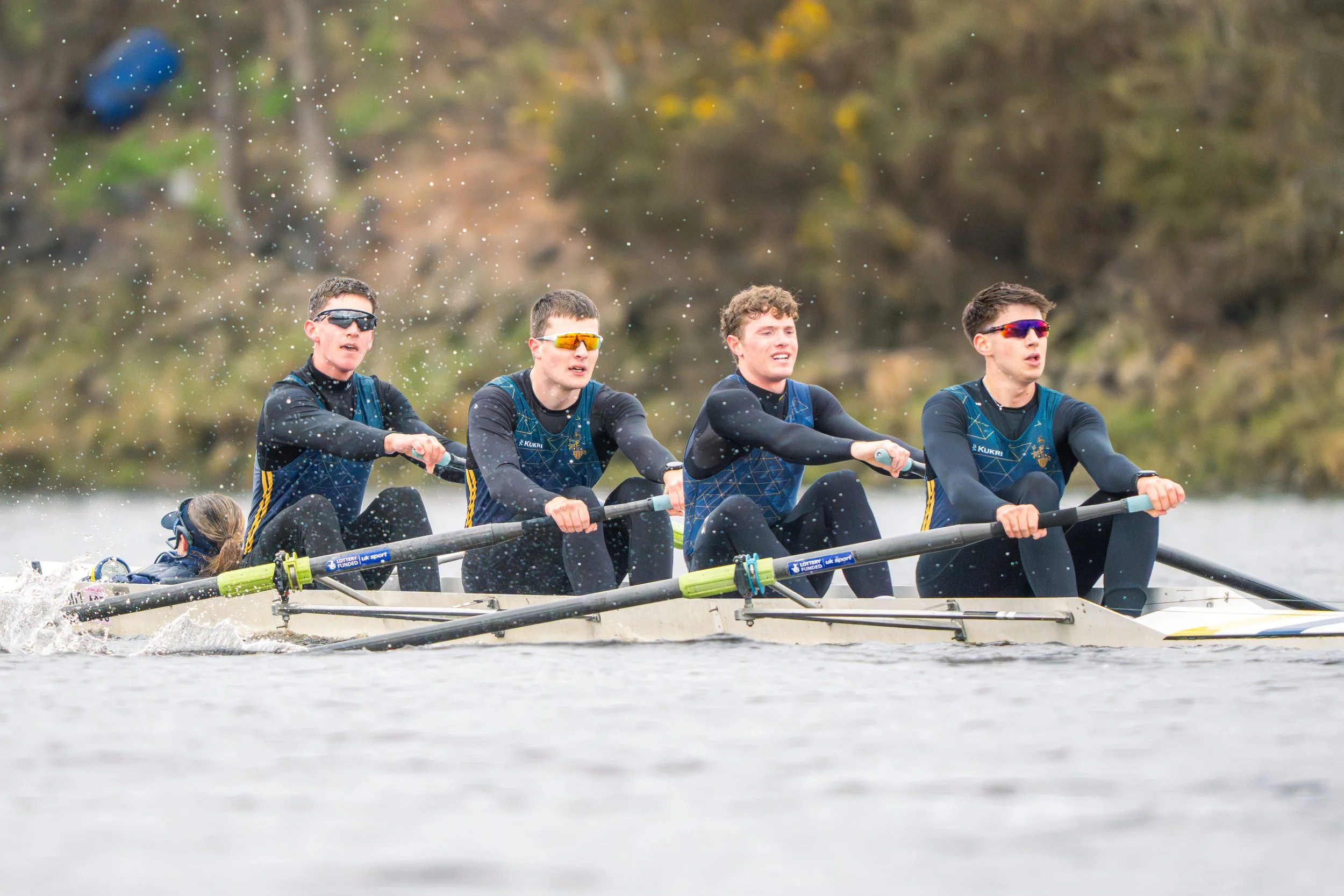 Four young men wearing sunglasses and matching athletic gear rowing a boat in a race on a waterway with a blurred background of autumn trees.