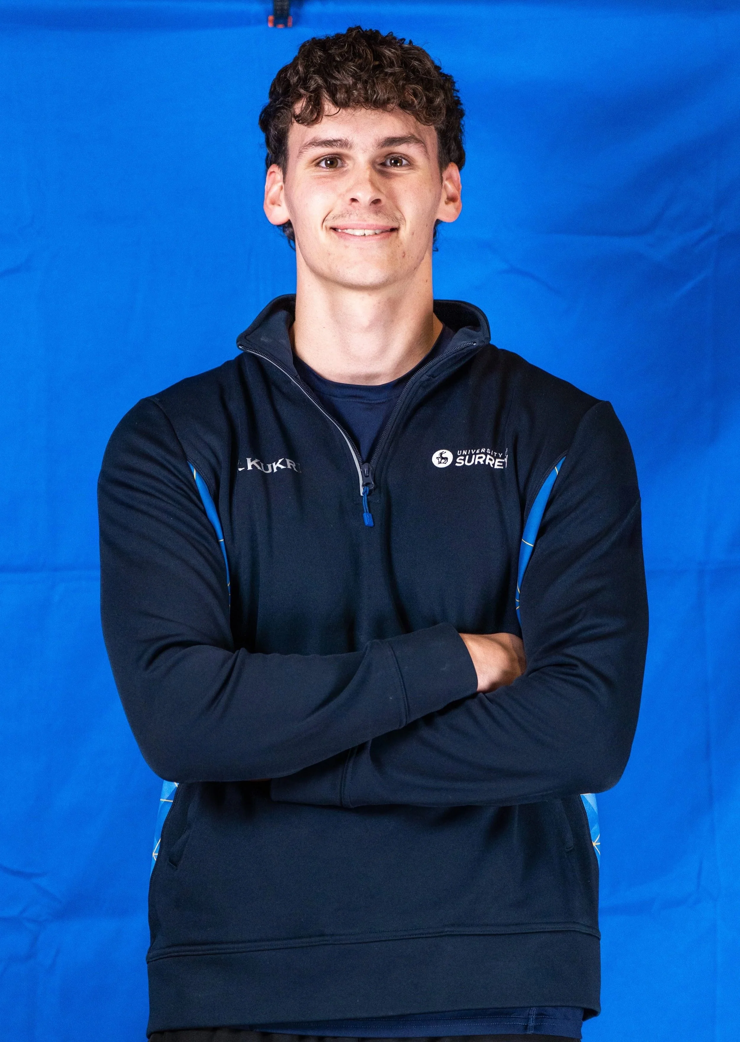 Young man with curly hair smiling and crossing arms, wearing a black sports jacket with logos, standing against a blue backdrop.