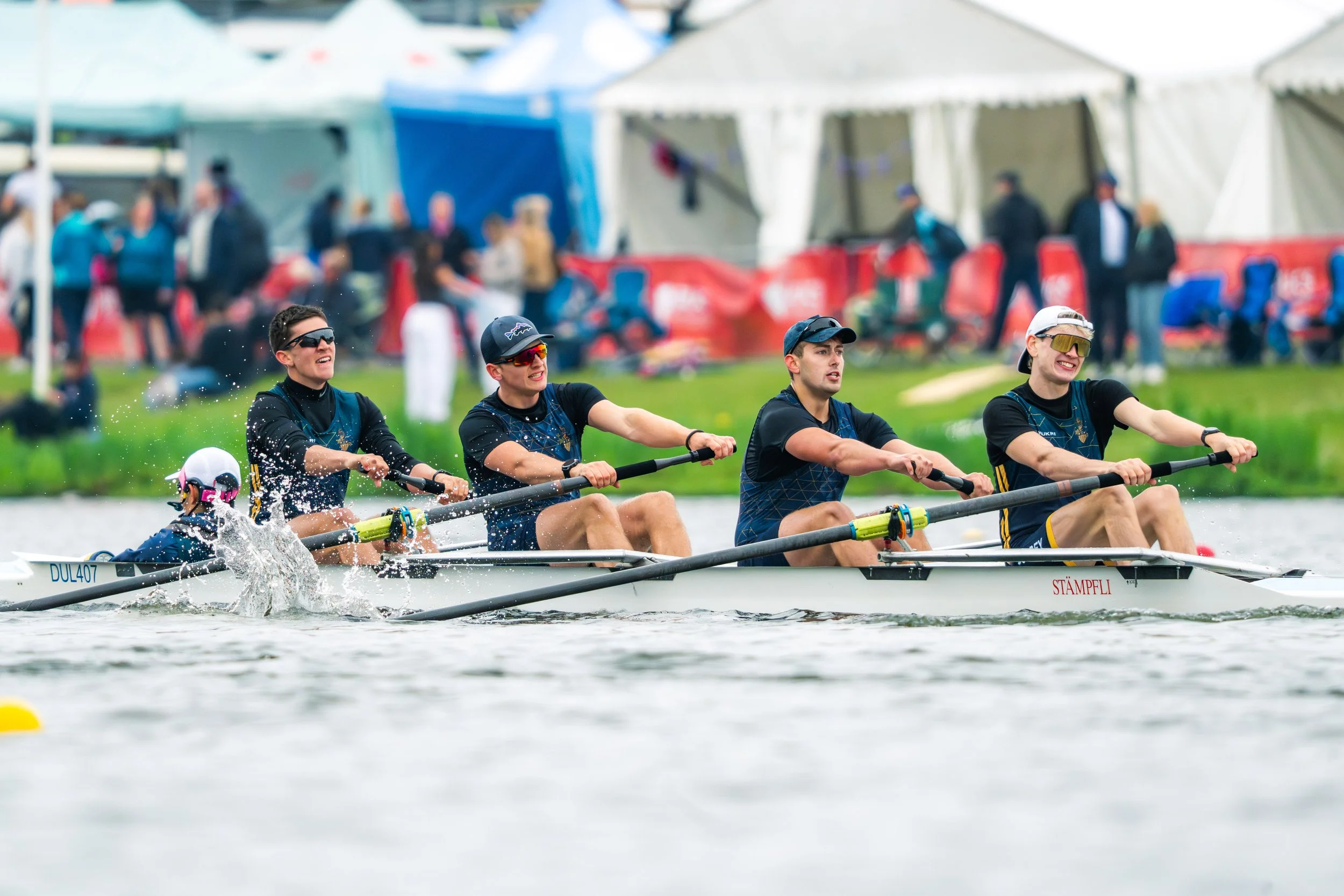 Four people in a rowing boat, competing in a race, with a crowd and tents in the background.