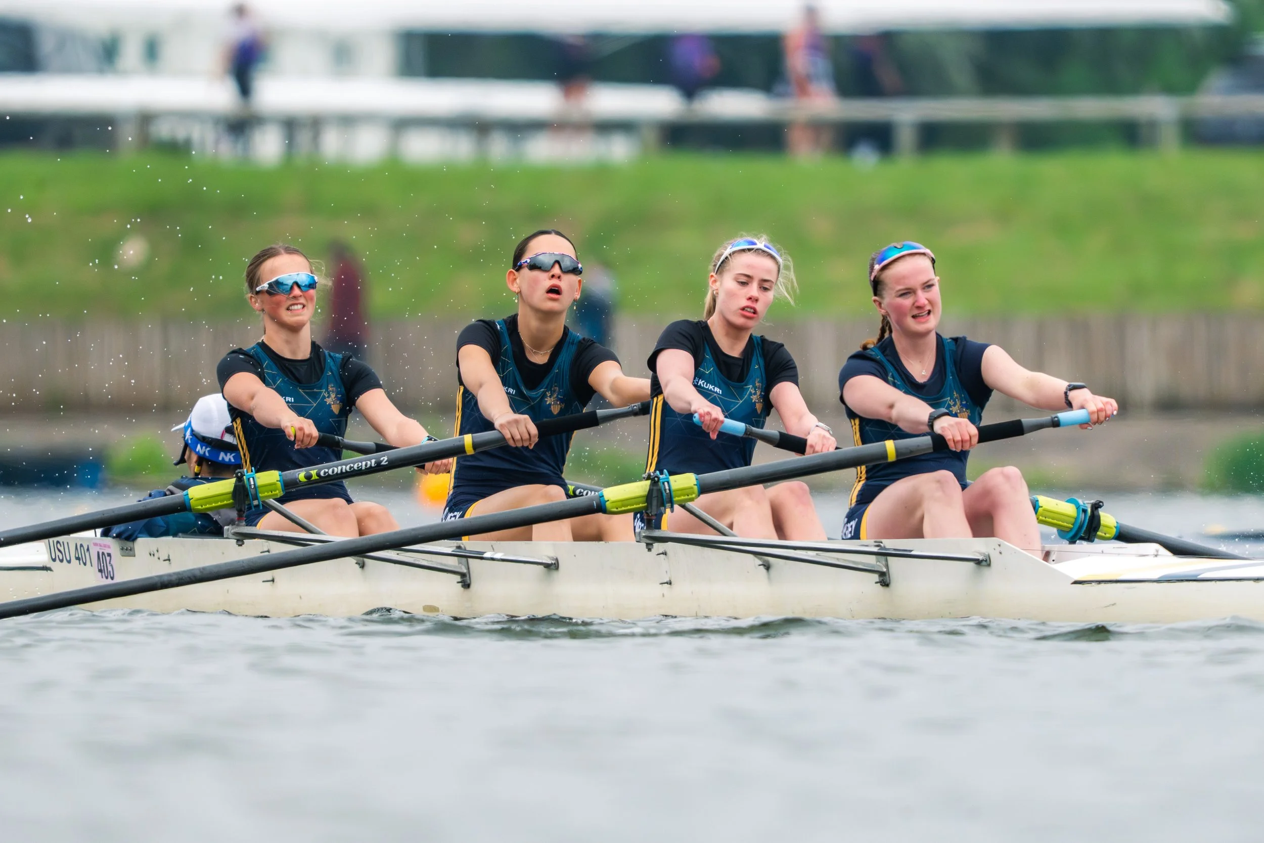 Four women in athletic gear and sunglasses are rowing a boat on a body of water, with a bridge and green landscape in the background.