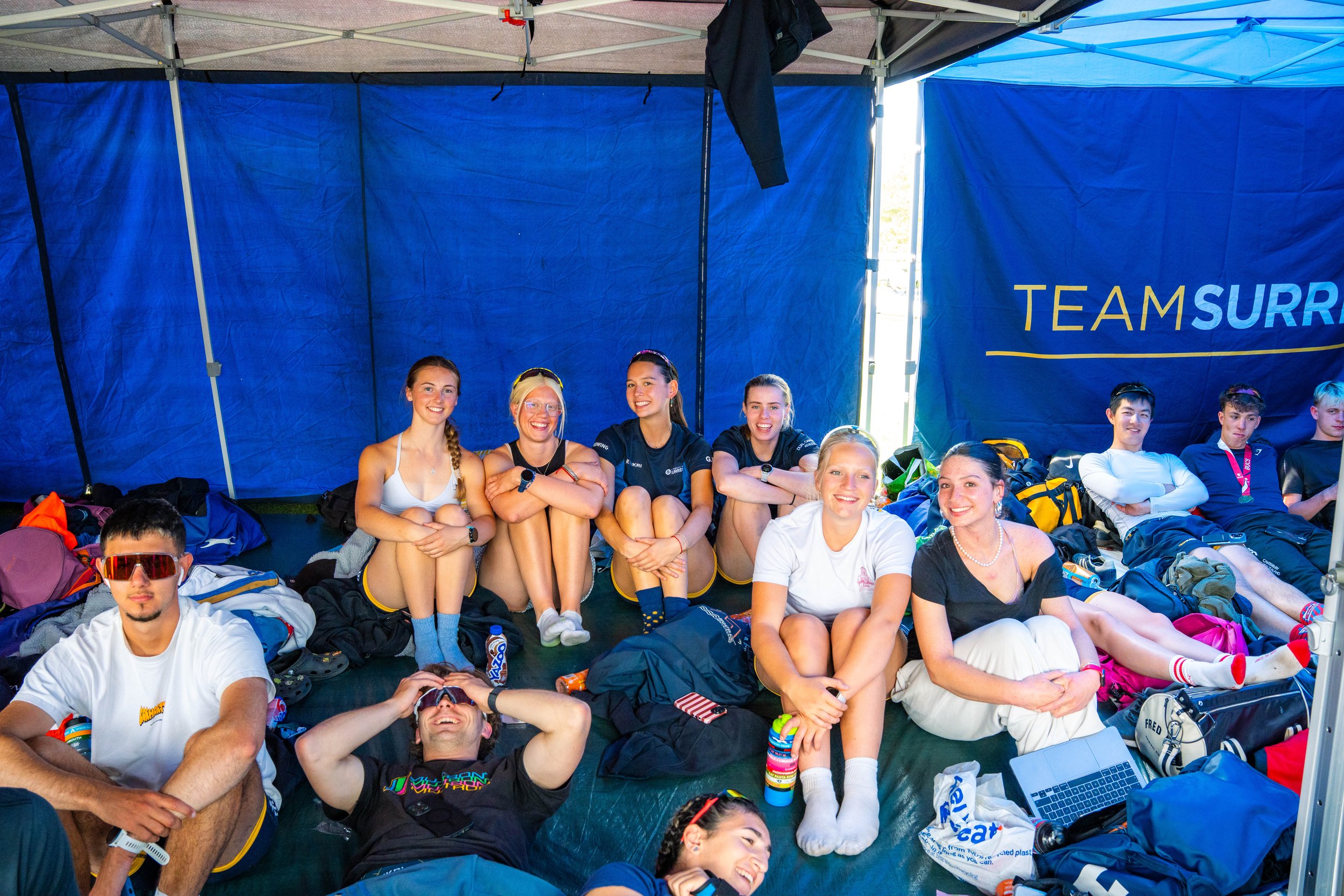 Group of young people sitting and lying inside a blue tent, smiling and relaxing. There are backpacks and bags around them, and a banner with the text "TEAM SURR" is visible behind them.