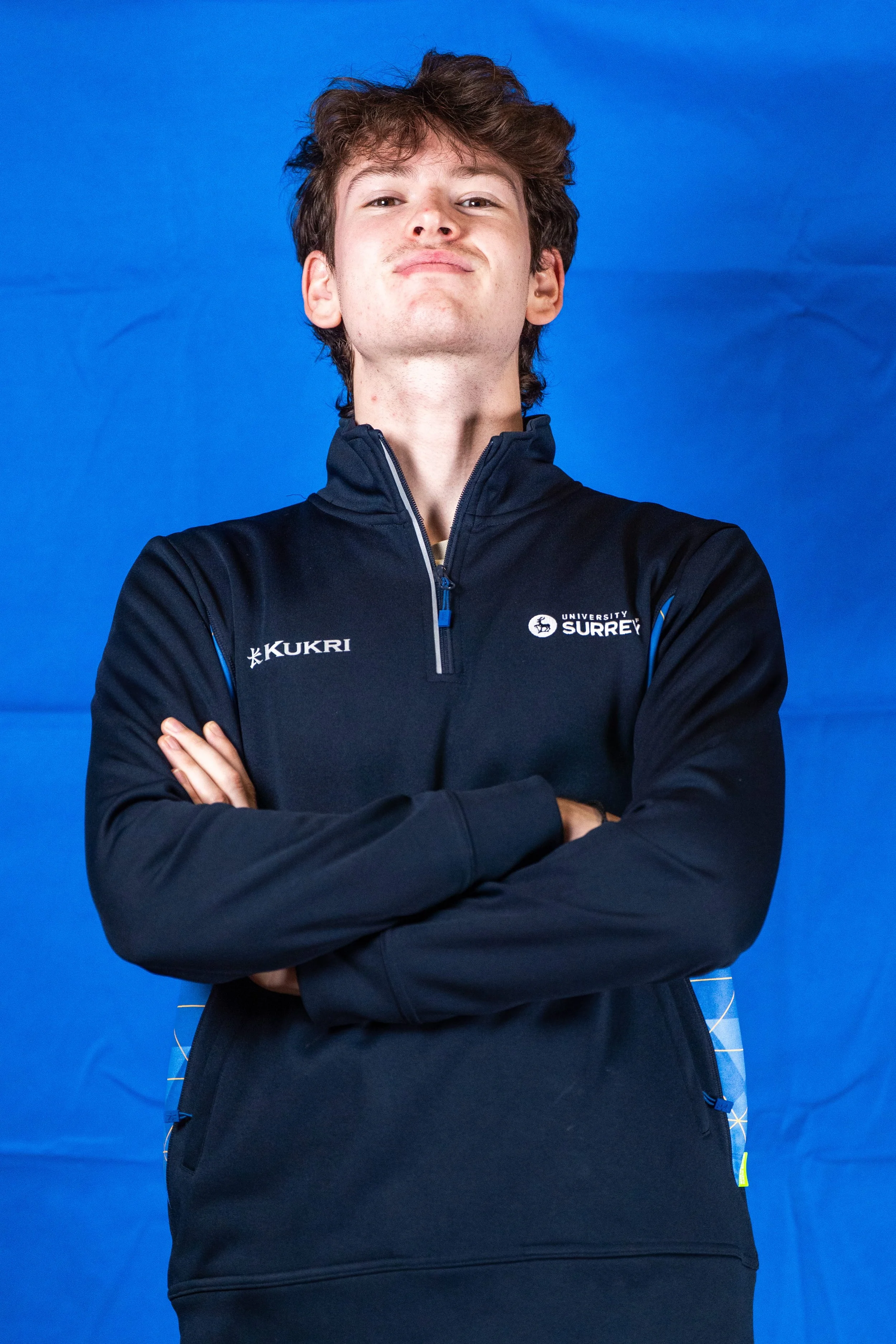 A young man with tousled brown hair and cross arms, wearing a black athletic jacket with University of Surrey logo, standing against a blue backdrop.