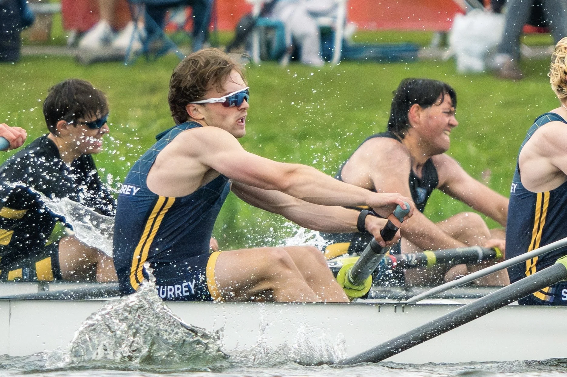A team of rowers in a boat racing on water, wearing dark blue athletic uniforms with yellow accents and sunglasses, with their hands gripping oars as they pull through the water, creating splashes and ripples.