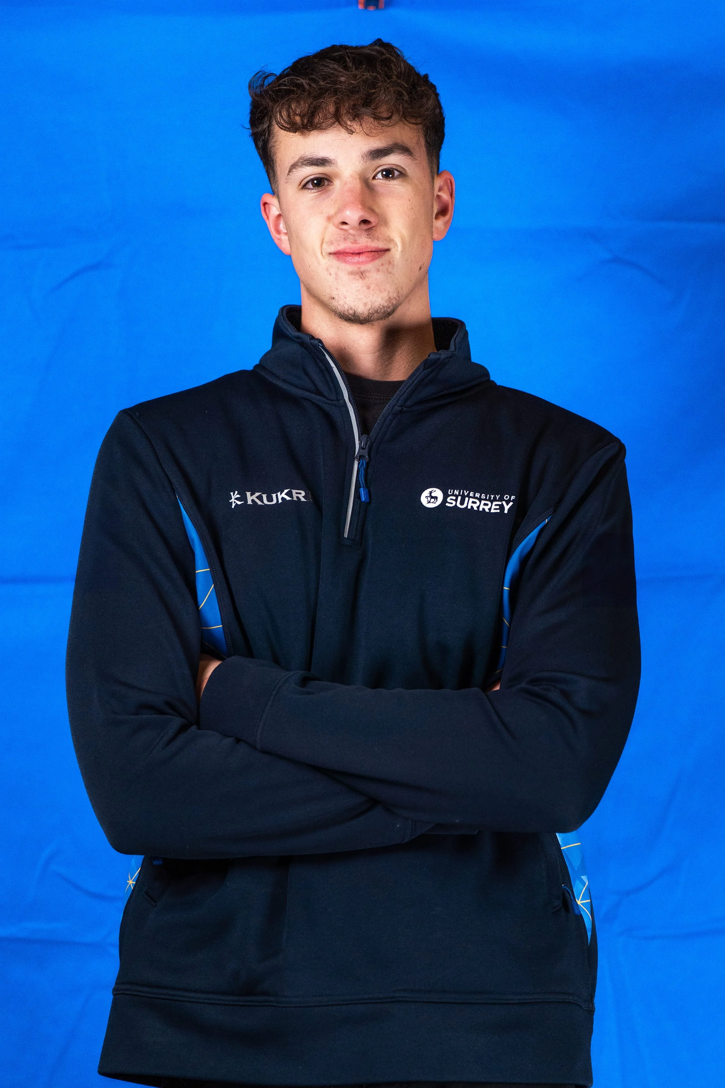 Young man with light skin, curly brown hair, and slight facial hair, wearing a black and blue University of Surrey jacket, standing with arms crossed in front of a blue background.