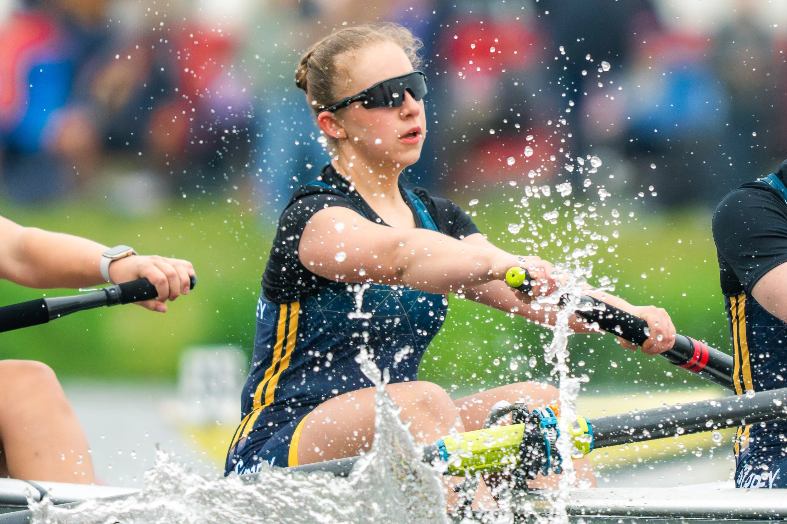 Young girl with blonde hair in a braid wearing sunglasses and a dark sports outfit, riding a watercraft and splashing water while participating in a water sport with other children in the background.