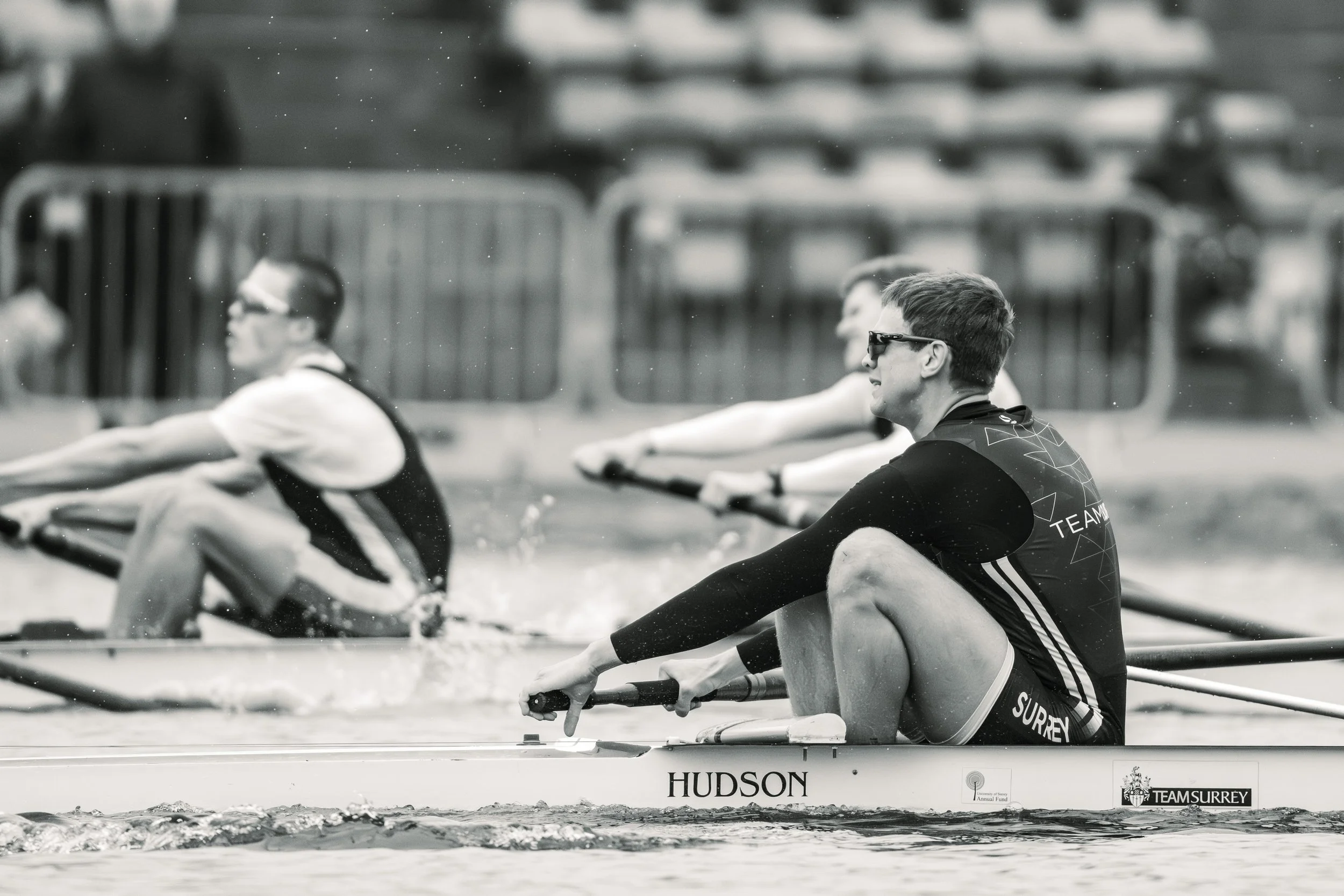 A black and white photograph of a group of rowers in a boat, dressed in athletic gear, rowing on a body of water with a blurred background of a fence and possibly other boats or structures. The boat has the words 'HUDSON' and 'TEAM SURREY' written on