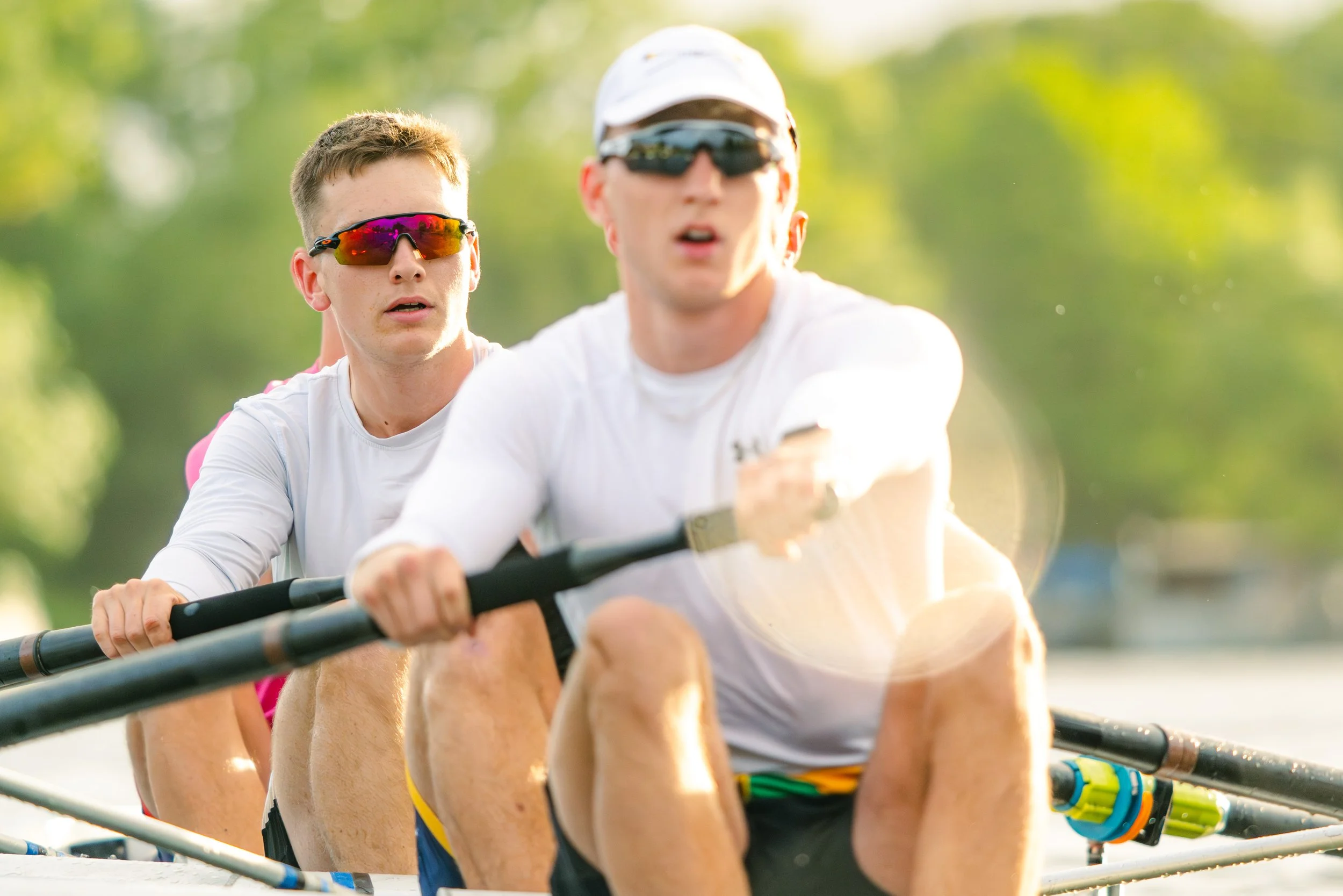 Four young men in a rowing boat competing in a race, all wearing sunglasses and athletic clothing, with a green blurred background of trees.