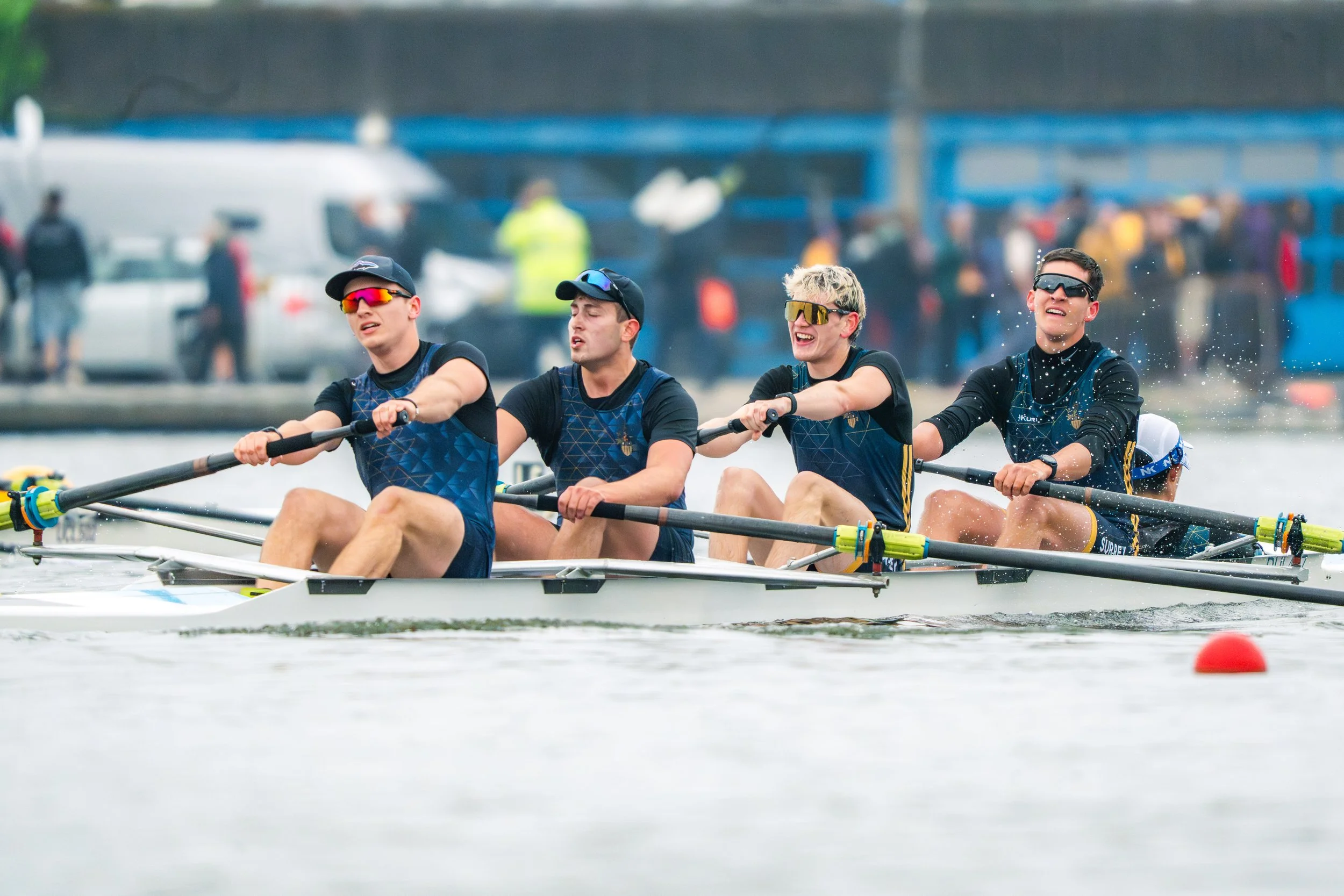 Four young men in a rowing boat on a body of water, wearing athletic gear and sunglasses, participating in a rowing competition or practice session with people and vehicles in the background.
