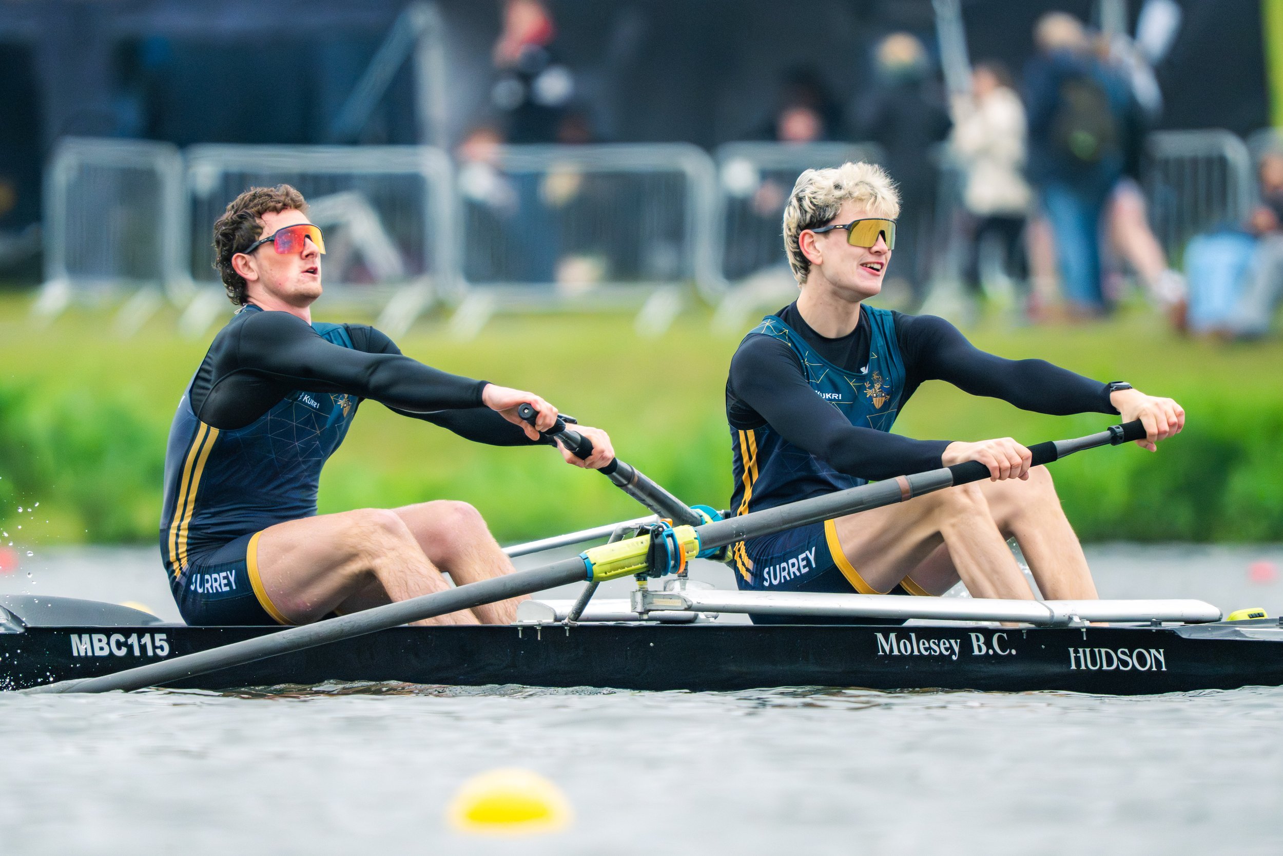 Two young men with sunglasses and athletic clothing are paddling a double scull rowing boat on a body of water, with a blurred background of spectators and barriers.