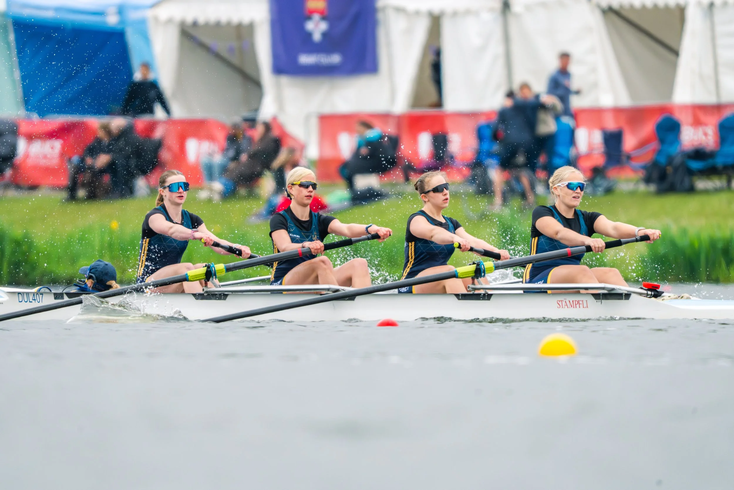 Four women wearing sunglasses and athletic outfits are competing in a rowing race on a body of water, pulling oars in unison. In the background, there are spectators, tents, and banners at the event.