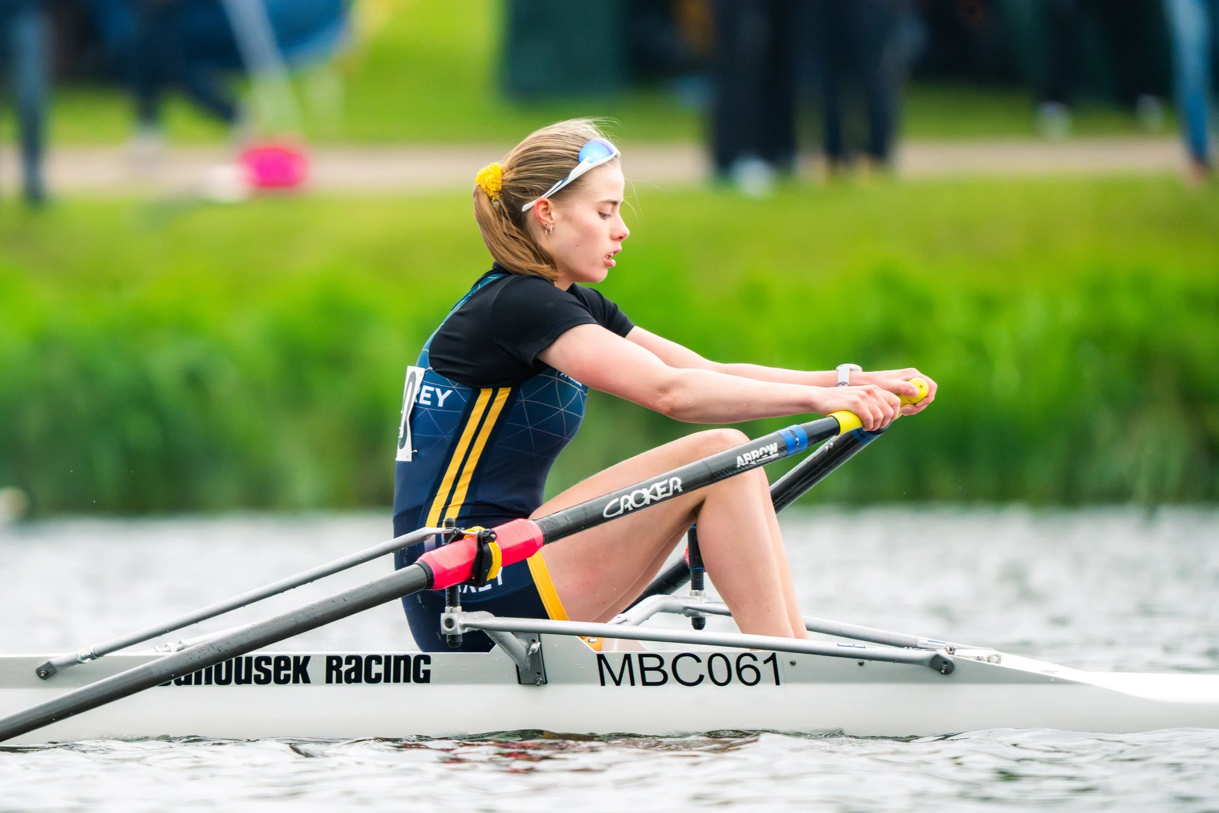 A young woman in athletic gear rowing on a river, with a focused expression, wearing sunglasses on her head, and a yellow flower hair accessory.