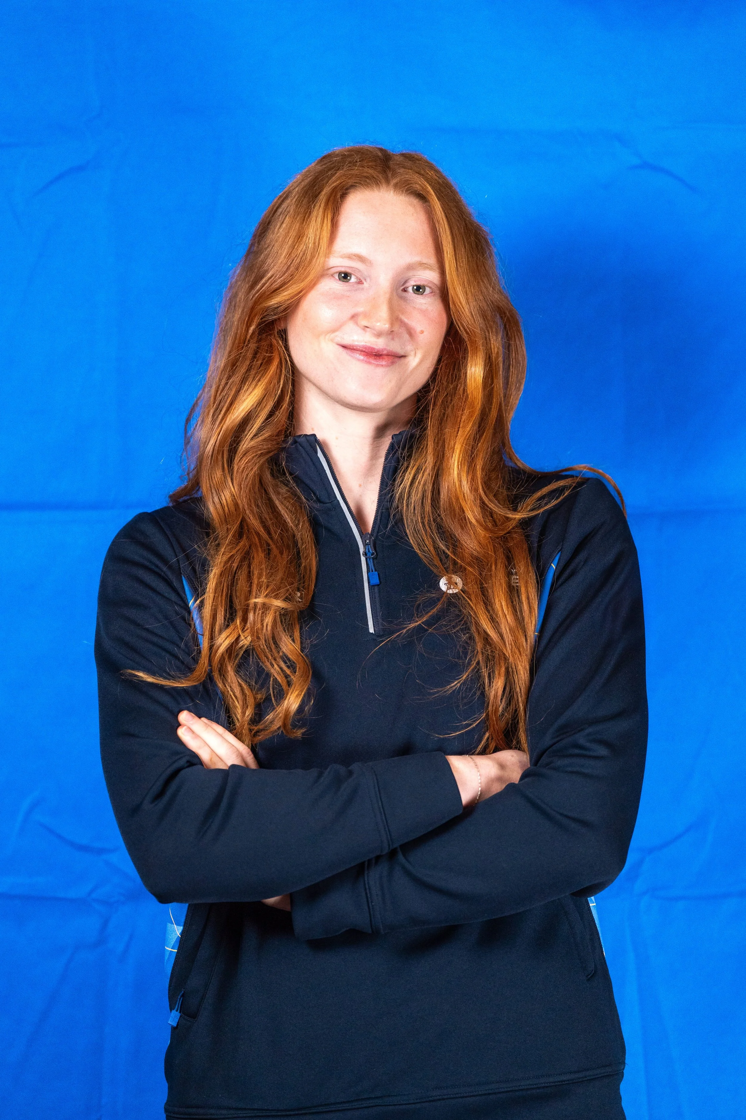 A woman with long red hair and a friendly smile, wearing a black athletic jacket with blue accents, standing with her arms crossed against a blue backdrop.