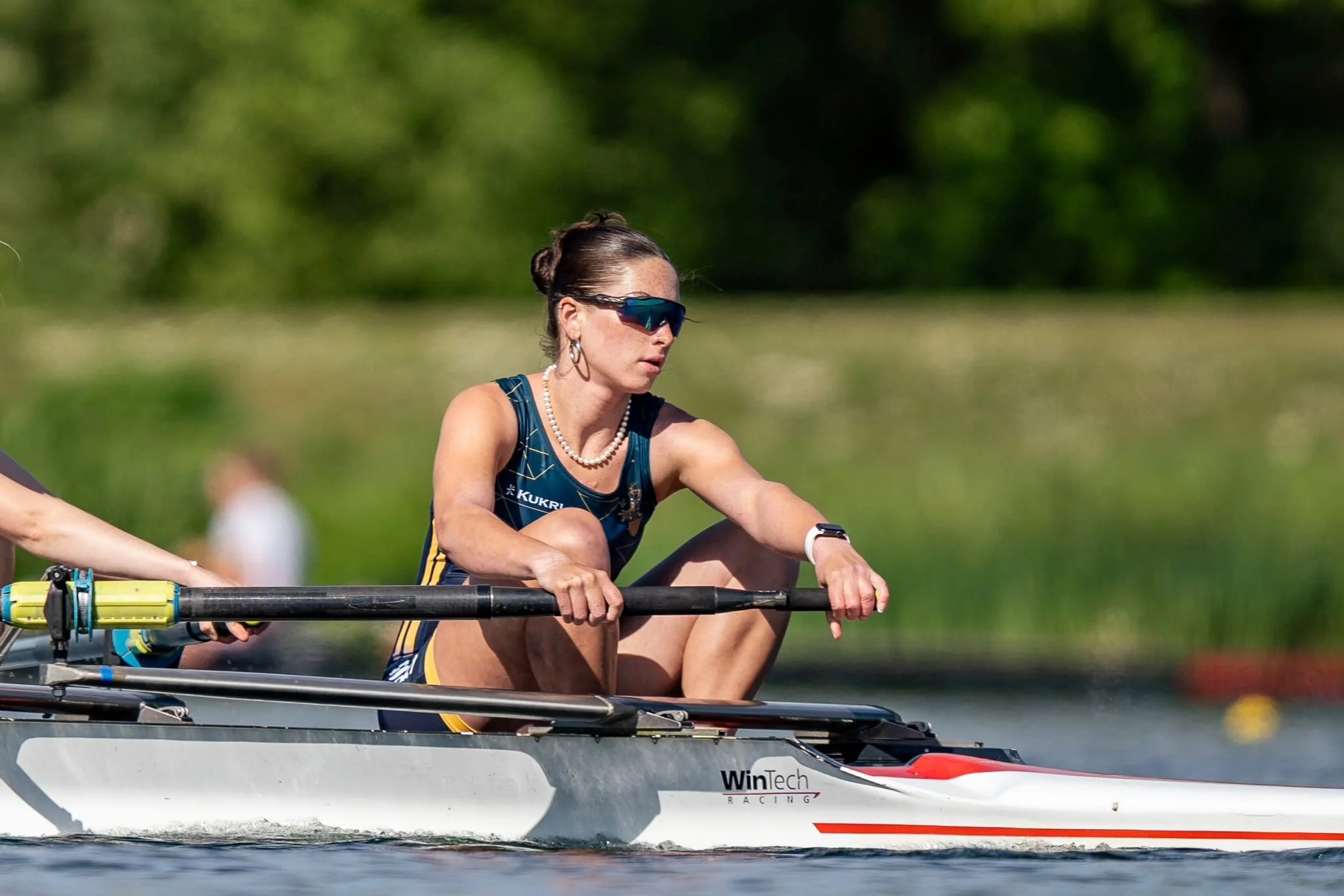 A woman is rowing in a racing shell on a body of water during daylight, wearing sunglasses and athletic gear, with greenery in the background.