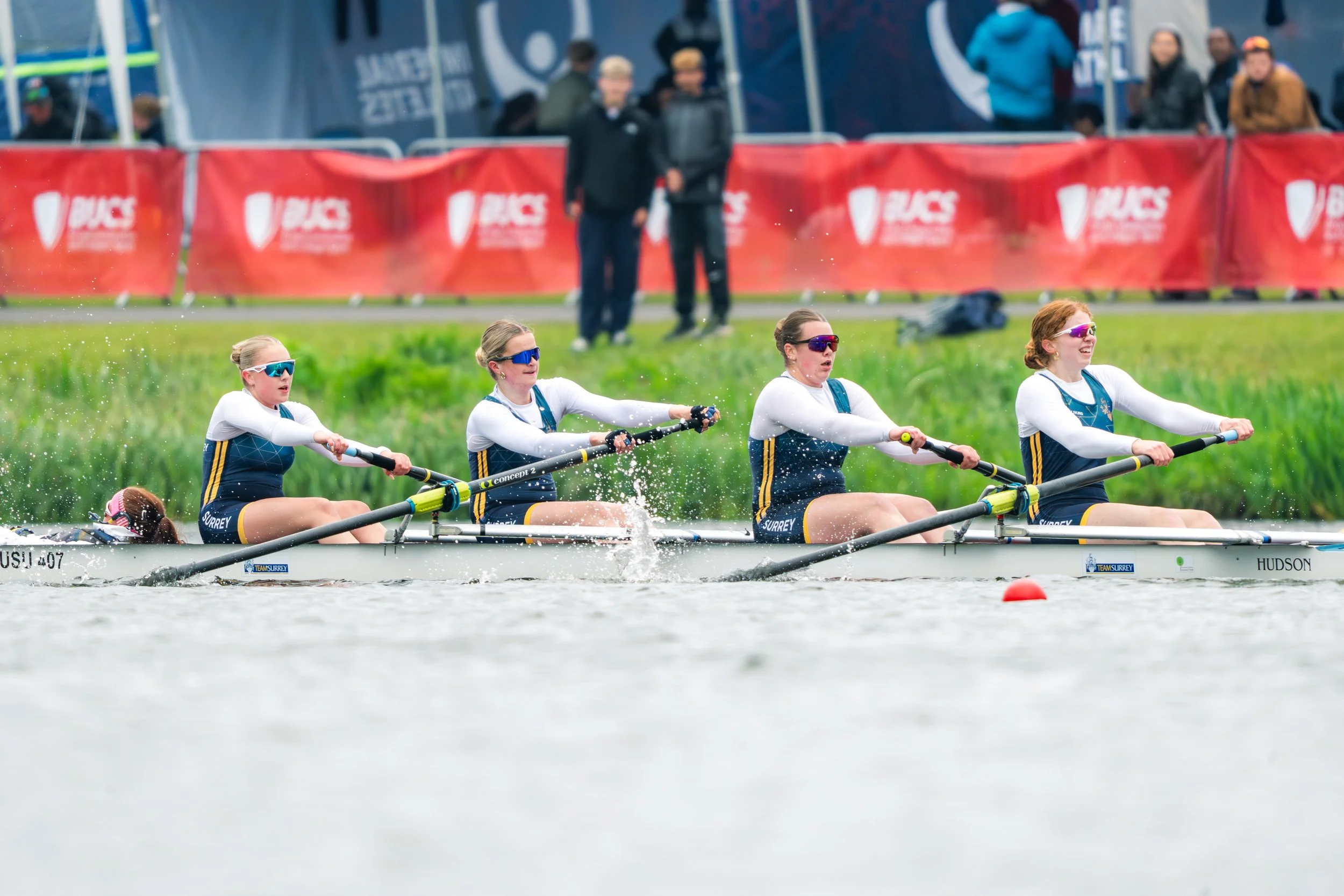 Four women in rowing uniforms and sunglasses competing in a rowing race on a river, with spectators watching behind a red banner.