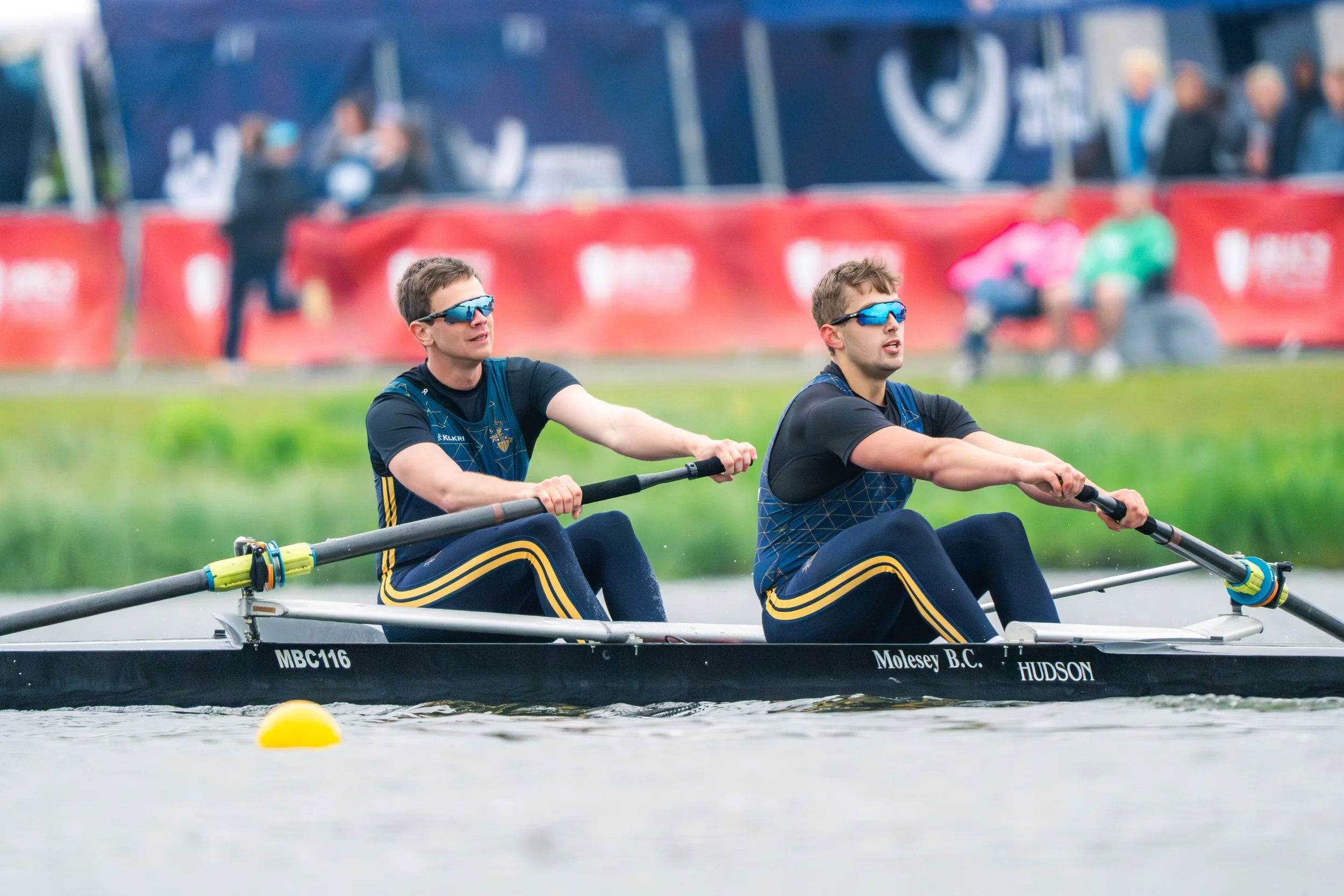 Two male athletes with sunglasses rowing a boat on a waterway during a race, with spectators and banners in the background.
