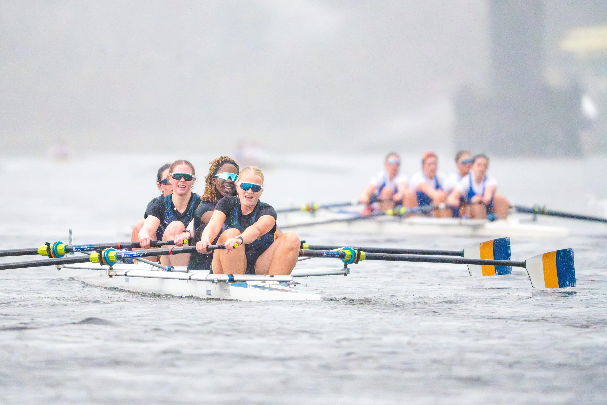 Women athletes competing in a rowing race on a water course, wearing sunglasses and sports gear, with a foggy or cloudy background.