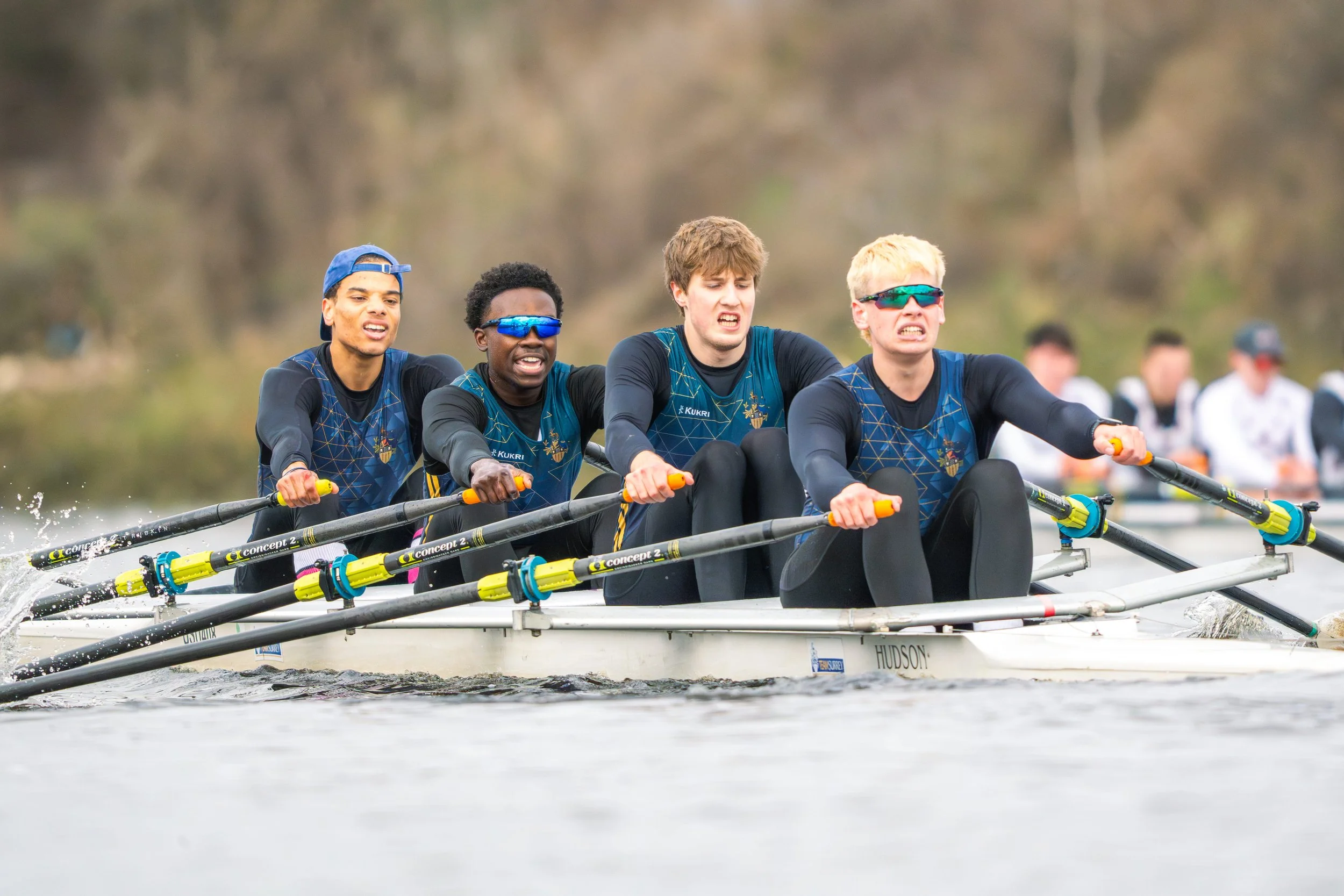 Four young male rowers in matching uniforms and sunglasses actively rowing a boat on a river during daytime.