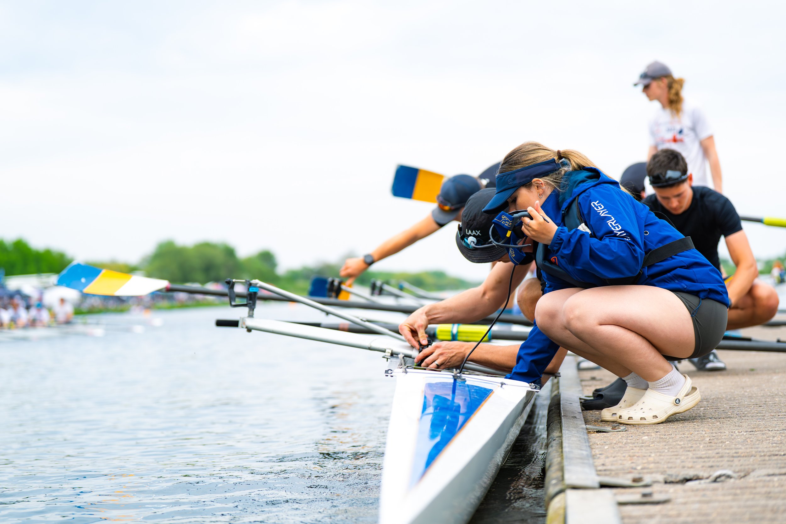 Rowers preparing their boats on a dock, crouched down and adjusting oars, with a background of water and sky.