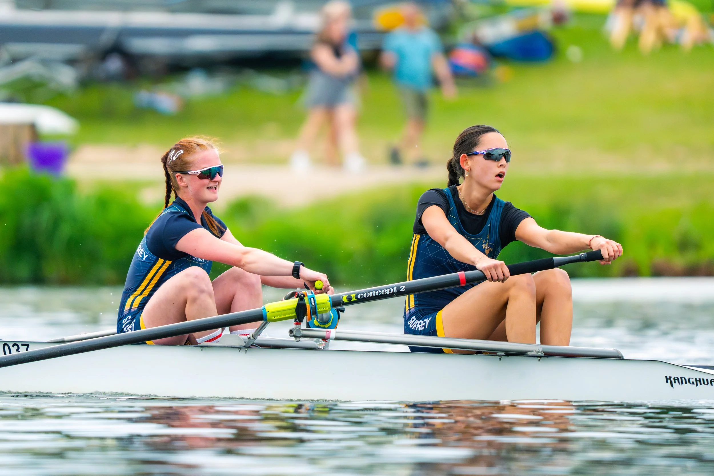 Two women wearing sunglasses and athletic gear are racing in a rowing boat on a body of water, with a grassy area and people in the background.