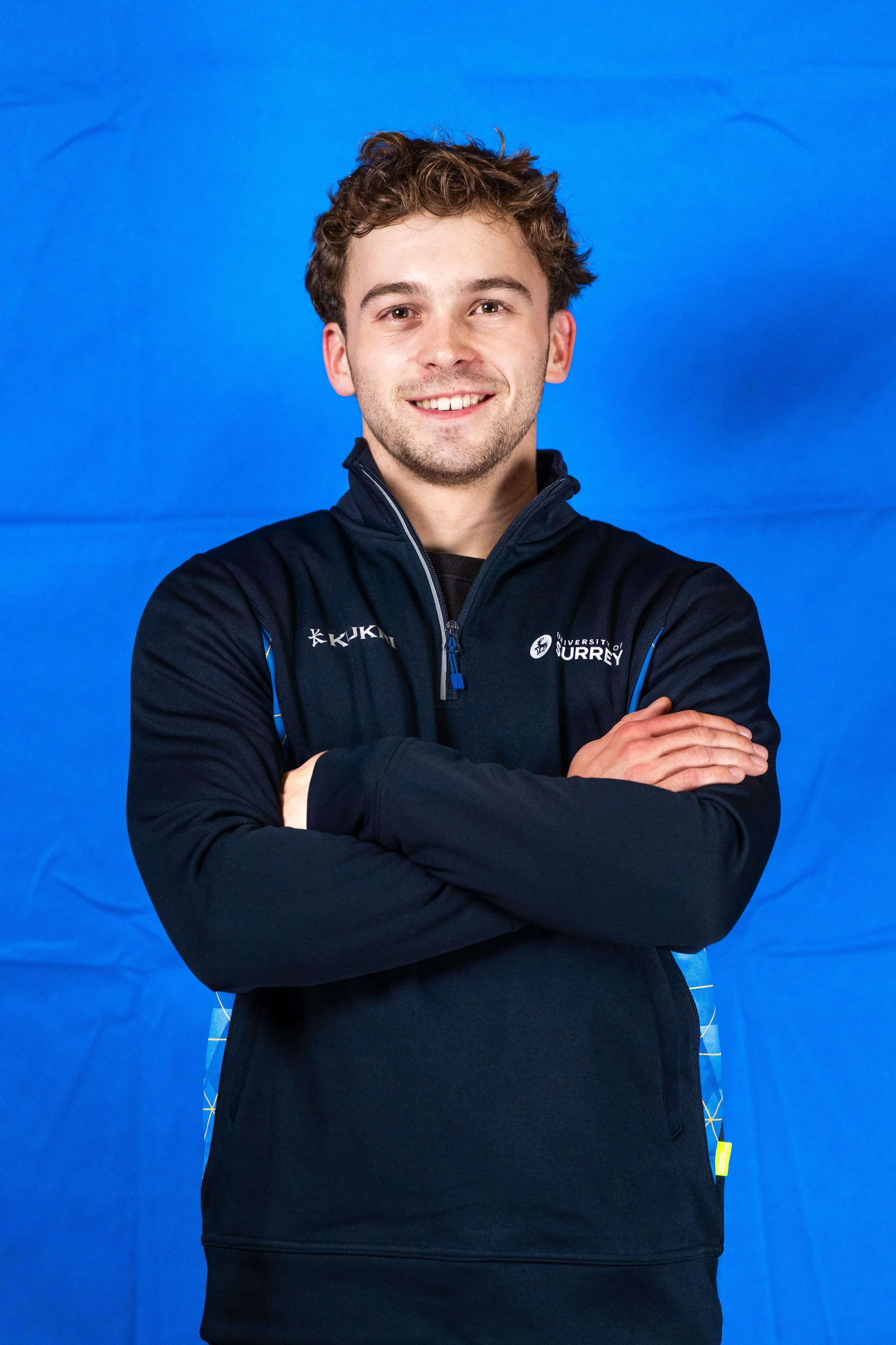 A young man with curly brown hair and light skin, smiling with arms crossed, wearing a black zip-up jacket with logos, posing against a bright blue background.