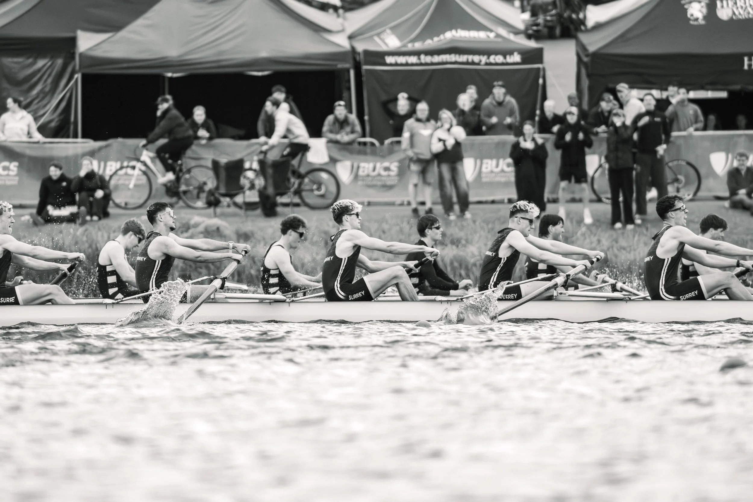 A team of rowers competing in a race on a body of water, with spectators and tents in the background.