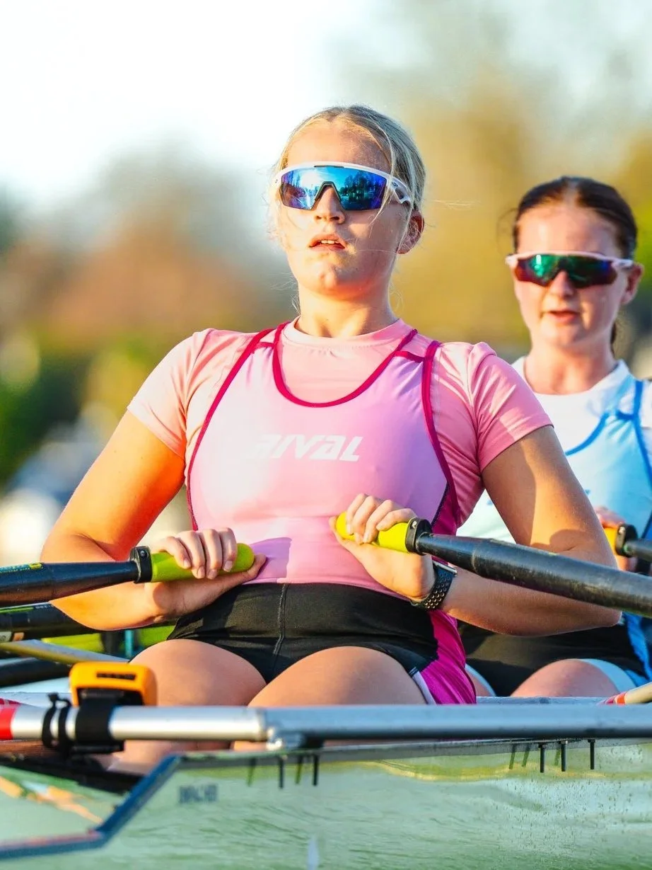 Two women rowing in a race on the water, wearing sunglasses and athletic clothing, with a blurred outdoor background.