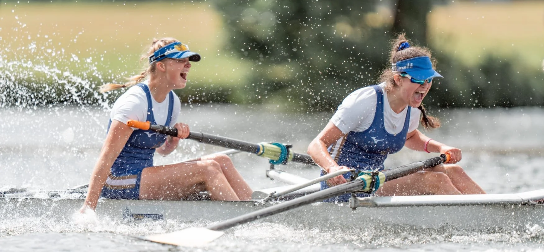 Two young women in rowing uniforms racing in a boat, splashing water, and experiencing intense effort and excitement.