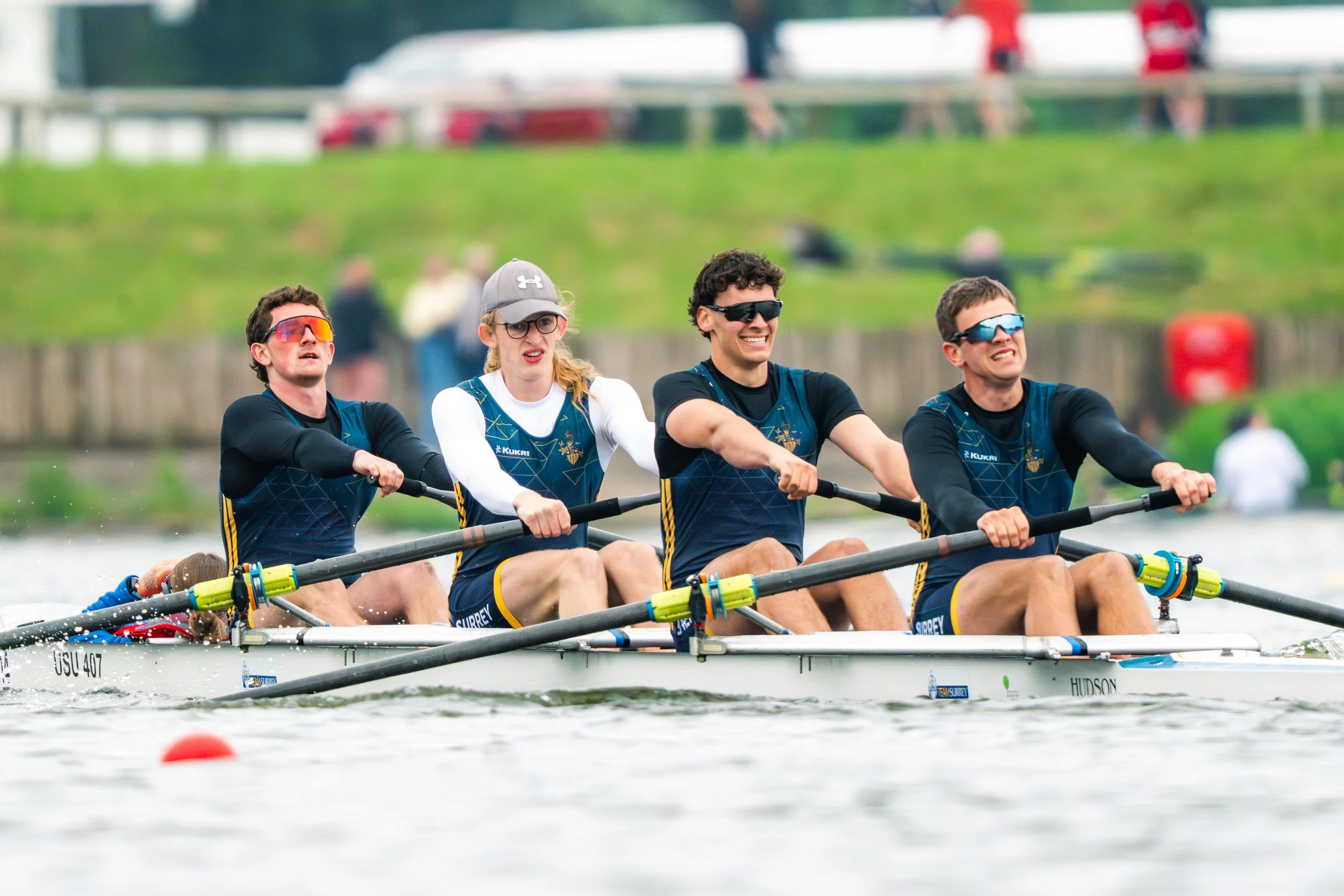 A team of four rowers wearing sunglasses and matching athletic uniforms, rowing a boat on a river during a sunny day.