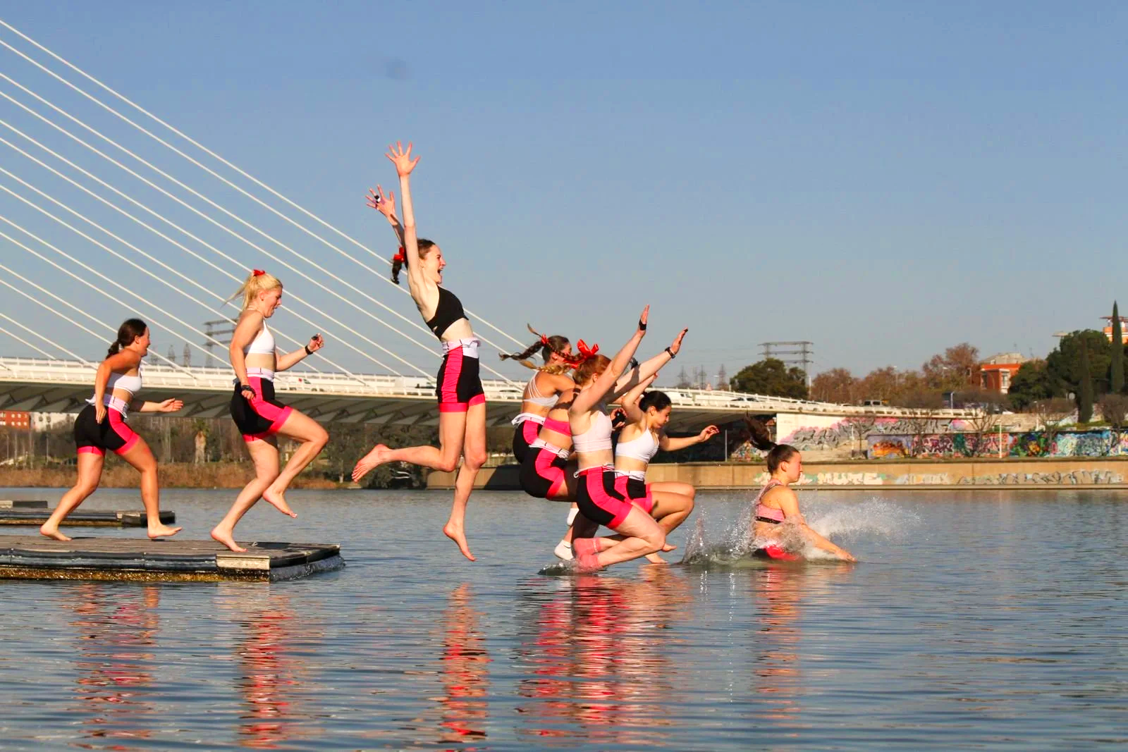 Eight young girls in athletic outfits jumping into or standing on a body of water beside a dock, with some landing in the water and others mid-air, during daytime. A bridge and cityscape are visible in the background.