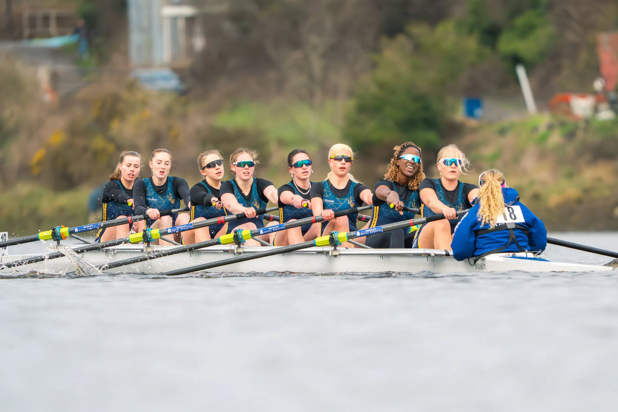 A team of female rowers competing in a rowing race on a river, wearing matching uniforms and sunglasses.