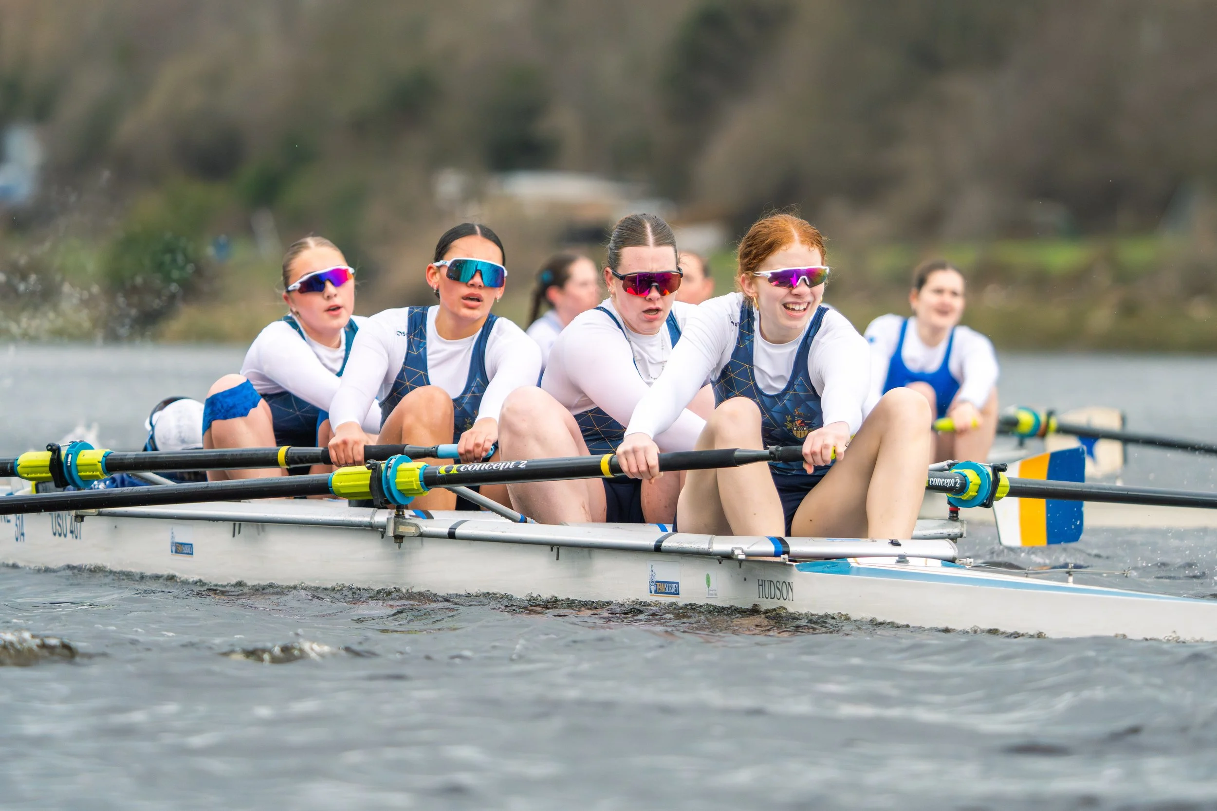 Women in a rowing boat competing in a race on a river, wearing sports gear and sunglasses, with a blurred natural background.