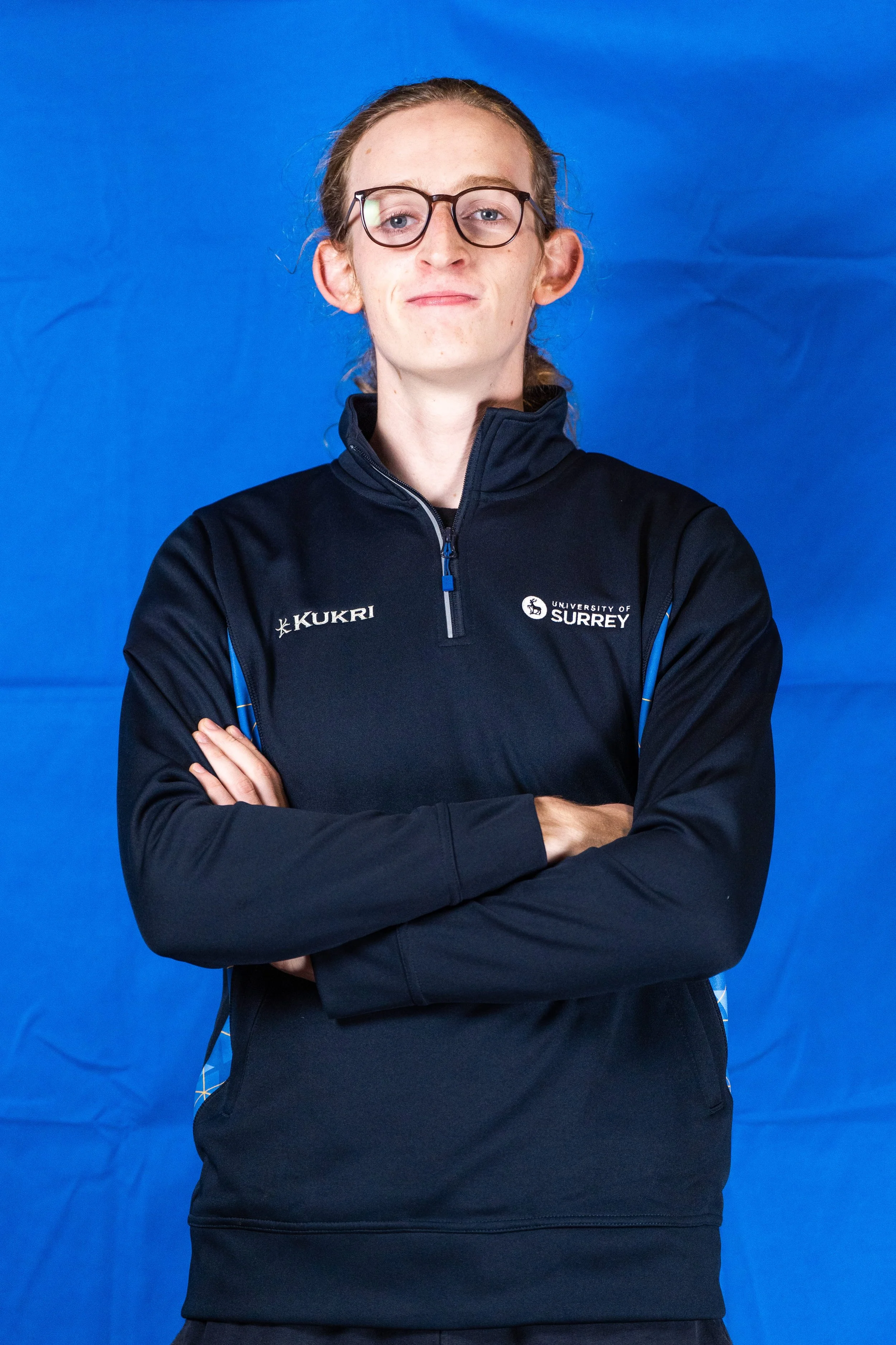 A young athlete with glasses standing with arms crossed in front of a blue background, wearing a black sports jacket with logos of Kukri and University of Surrey.