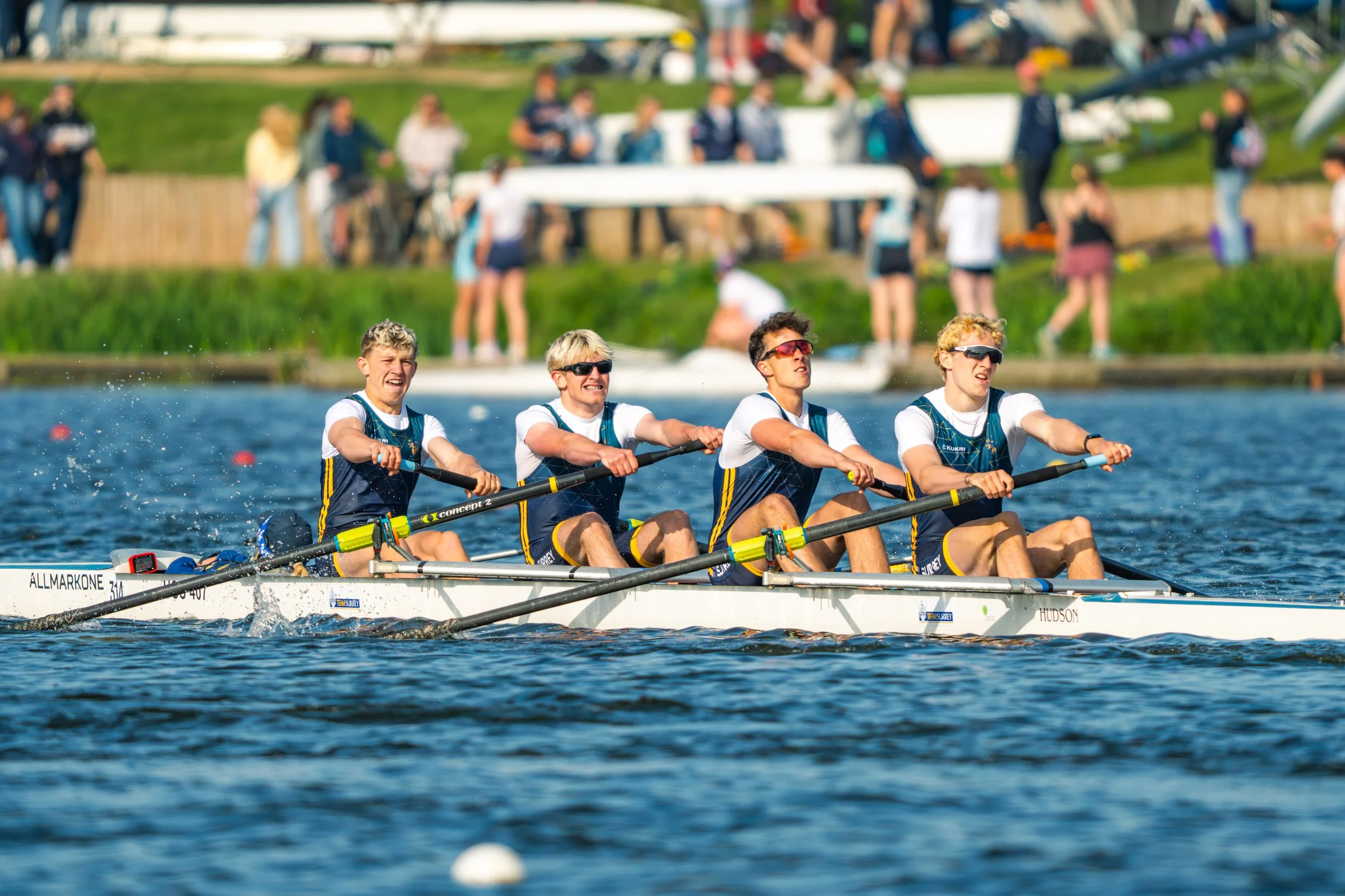 A team of four young men wearing white shirts and dark rowing uniforms paddling a boat in a competitive rowing race on a body of water, with spectators and greenery in the background.