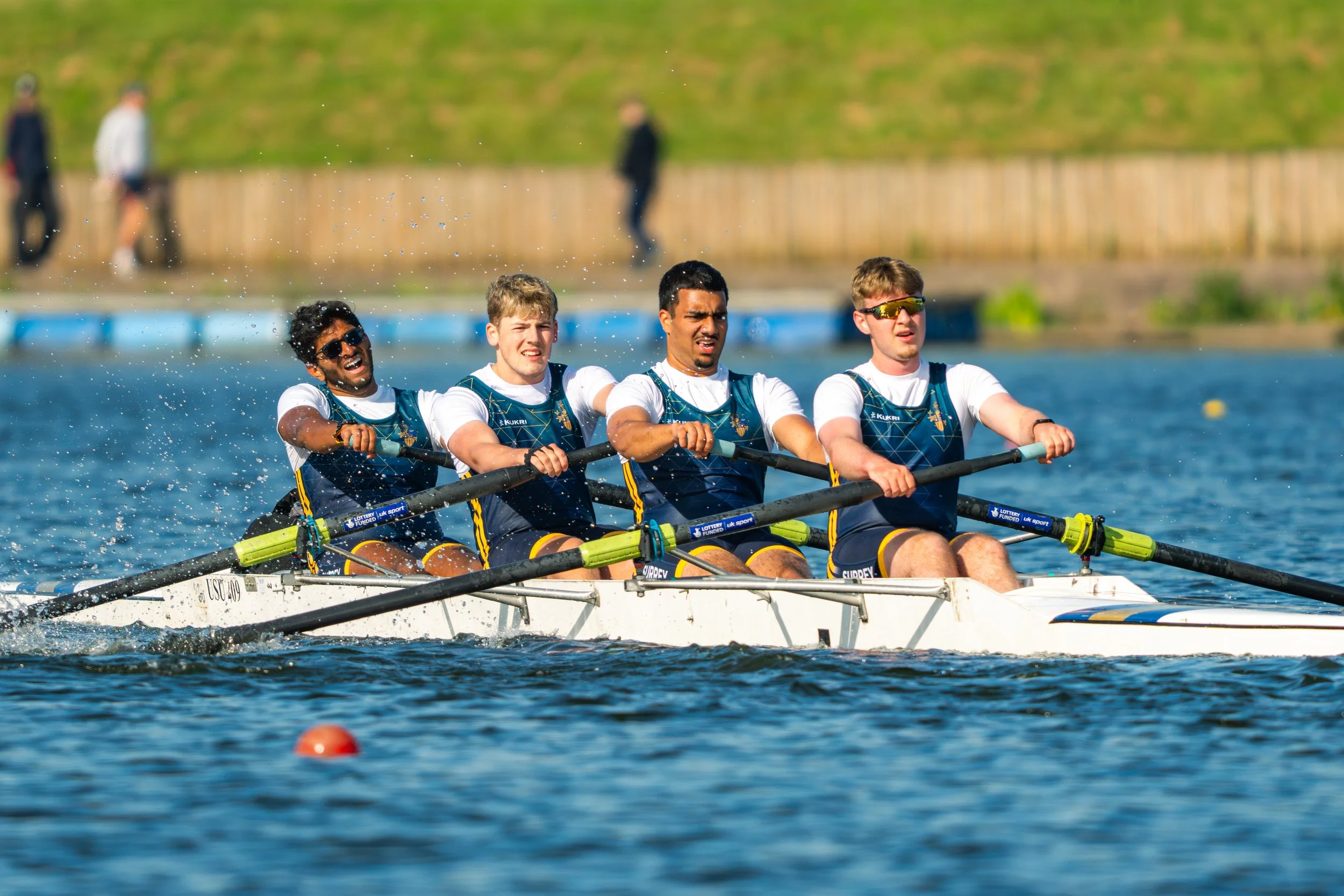 A team of four male rowers in a boat competing in a rowing race on a body of water, with a grassy bank and people walking in the background.