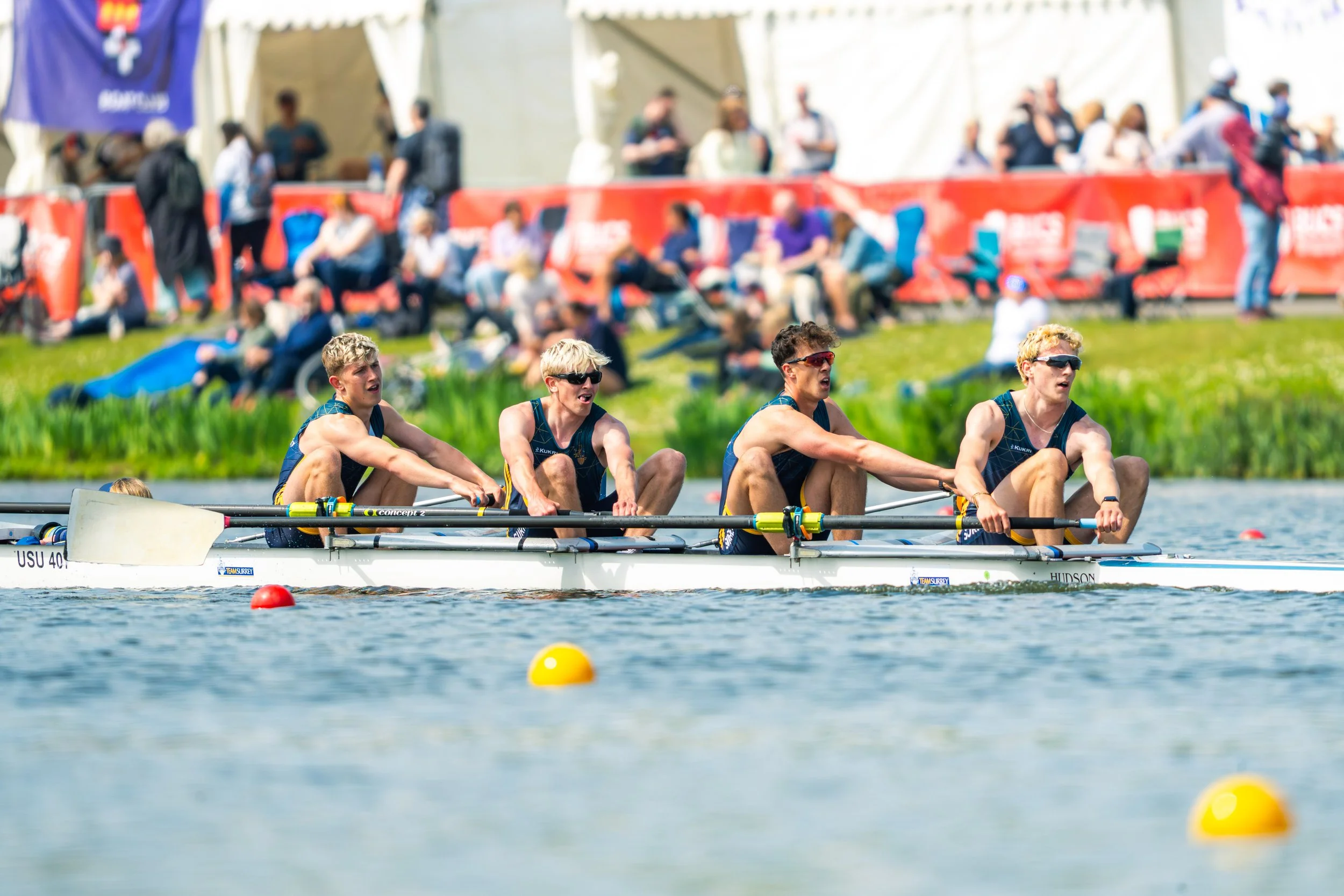 Four young male rowers wearing blue athletic uniforms and sunglasses, intensely rowing a boat on a lake during a race, with spectators standing behind a red barrier in the background.