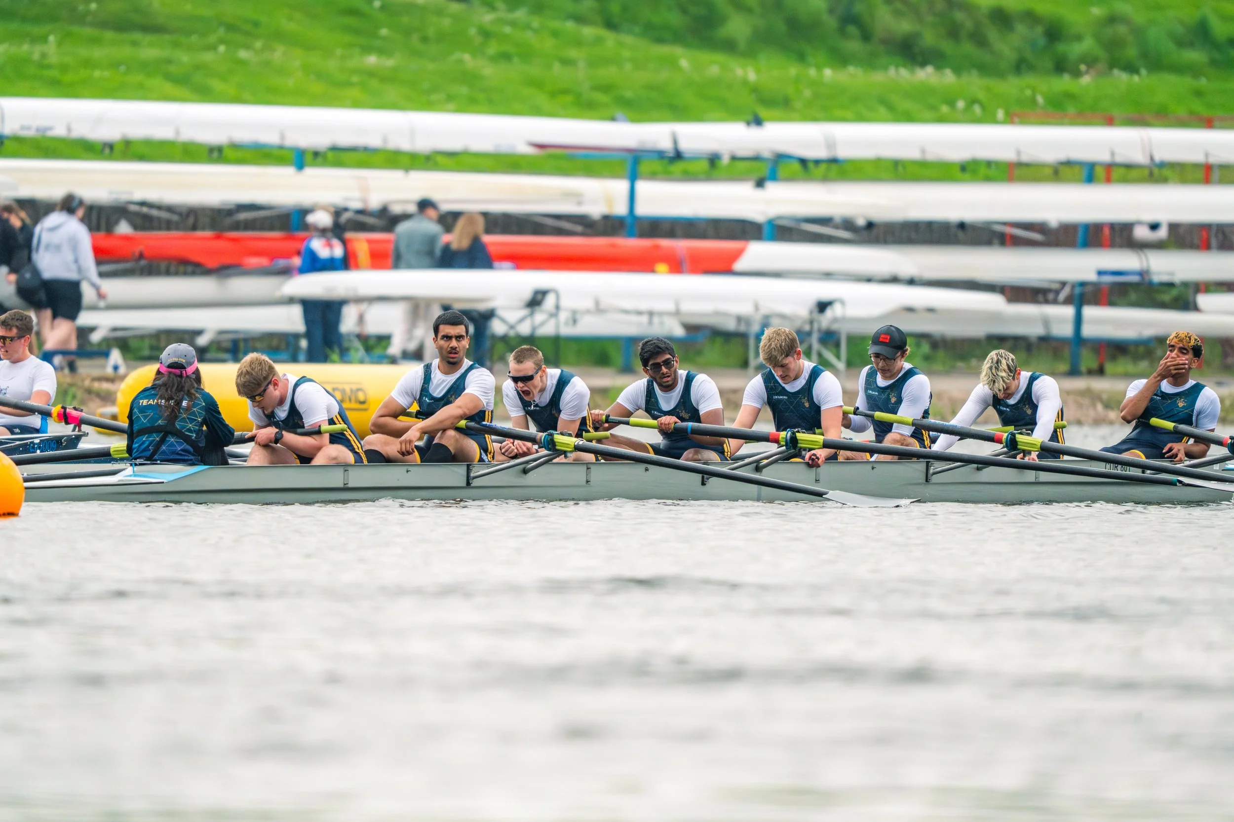 A group of rowers wearing matching uniforms sitting in a rowing shell on a river, with others walking on the shore and rowing shells stored on racks in the background.
