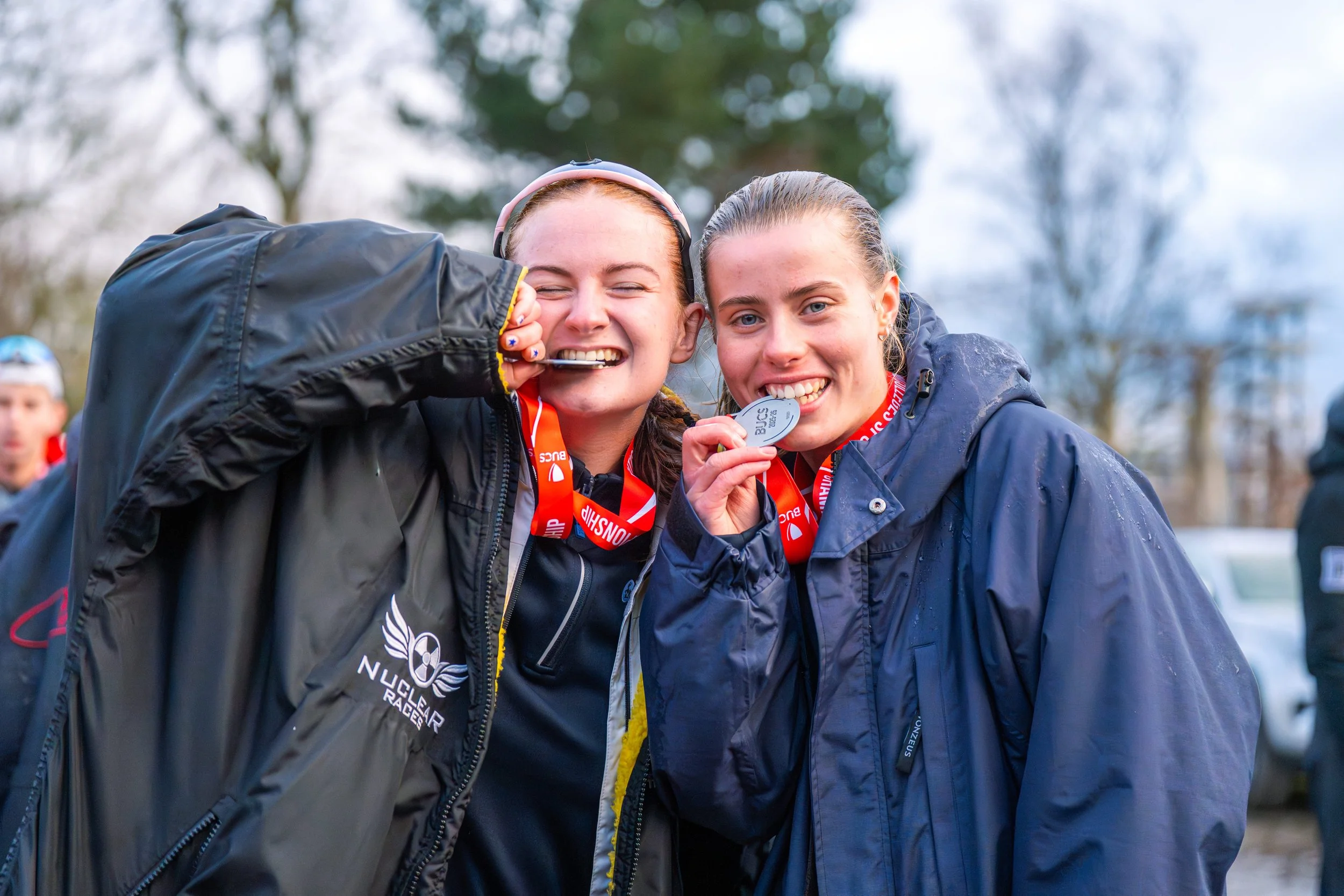 Two young women celebrating after a race, wearing medals around their necks, smiling and posing for the photo outdoors on a cloudy day.