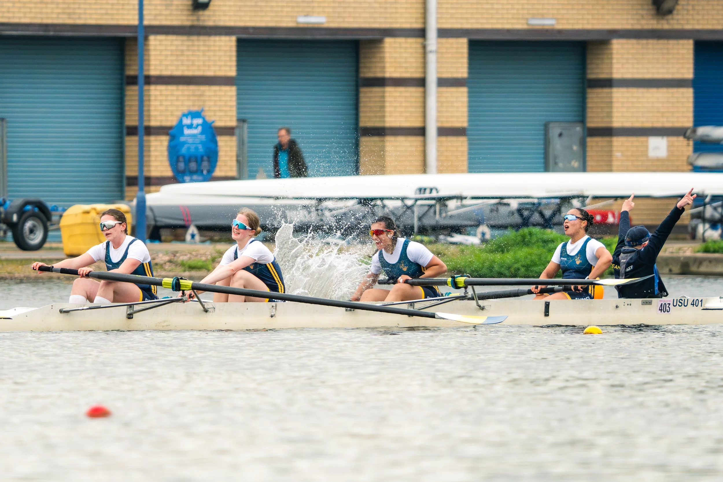 Women in a rowing boat racing on the water with a building and shoreline in the background
