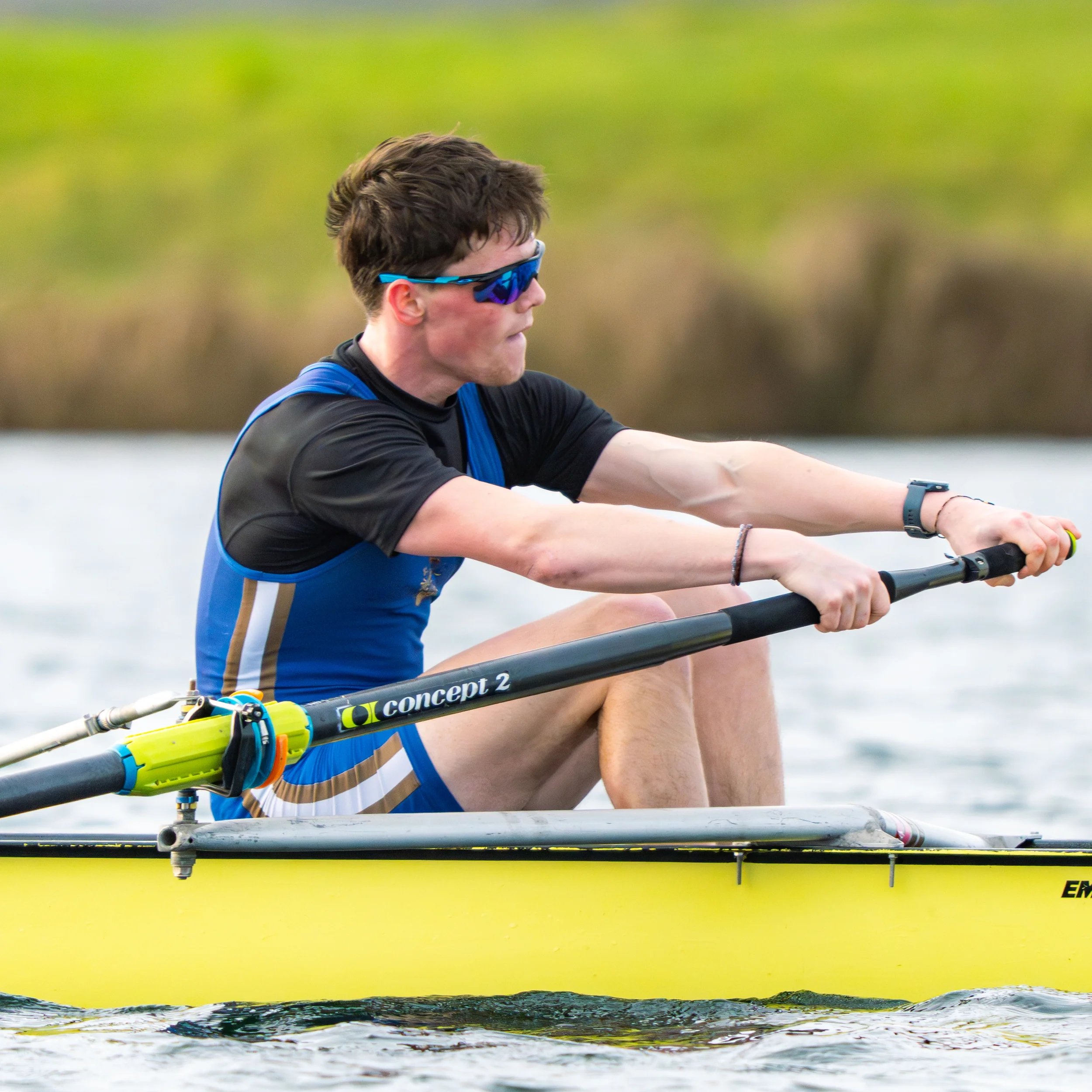 A young man with dark hair wearing sunglasses and a blue sports shirt paddles a kayak in a body of water, with a blurred green and yellow background.